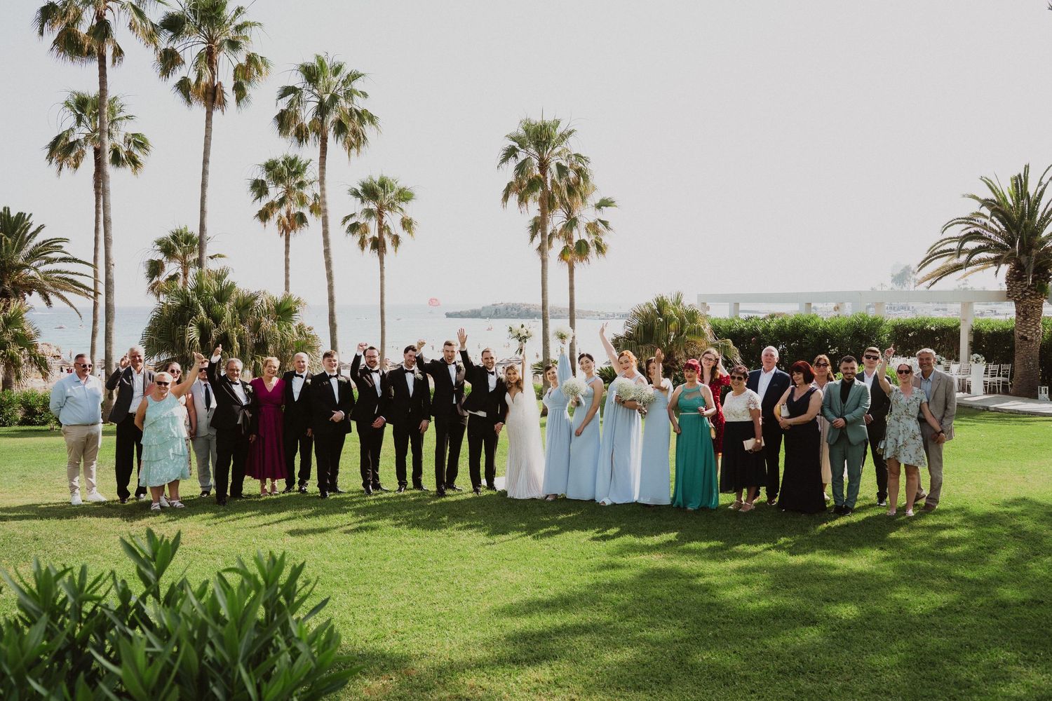 Large wedding party poses on lush green lawn with tall palm trees and ocean views in background.