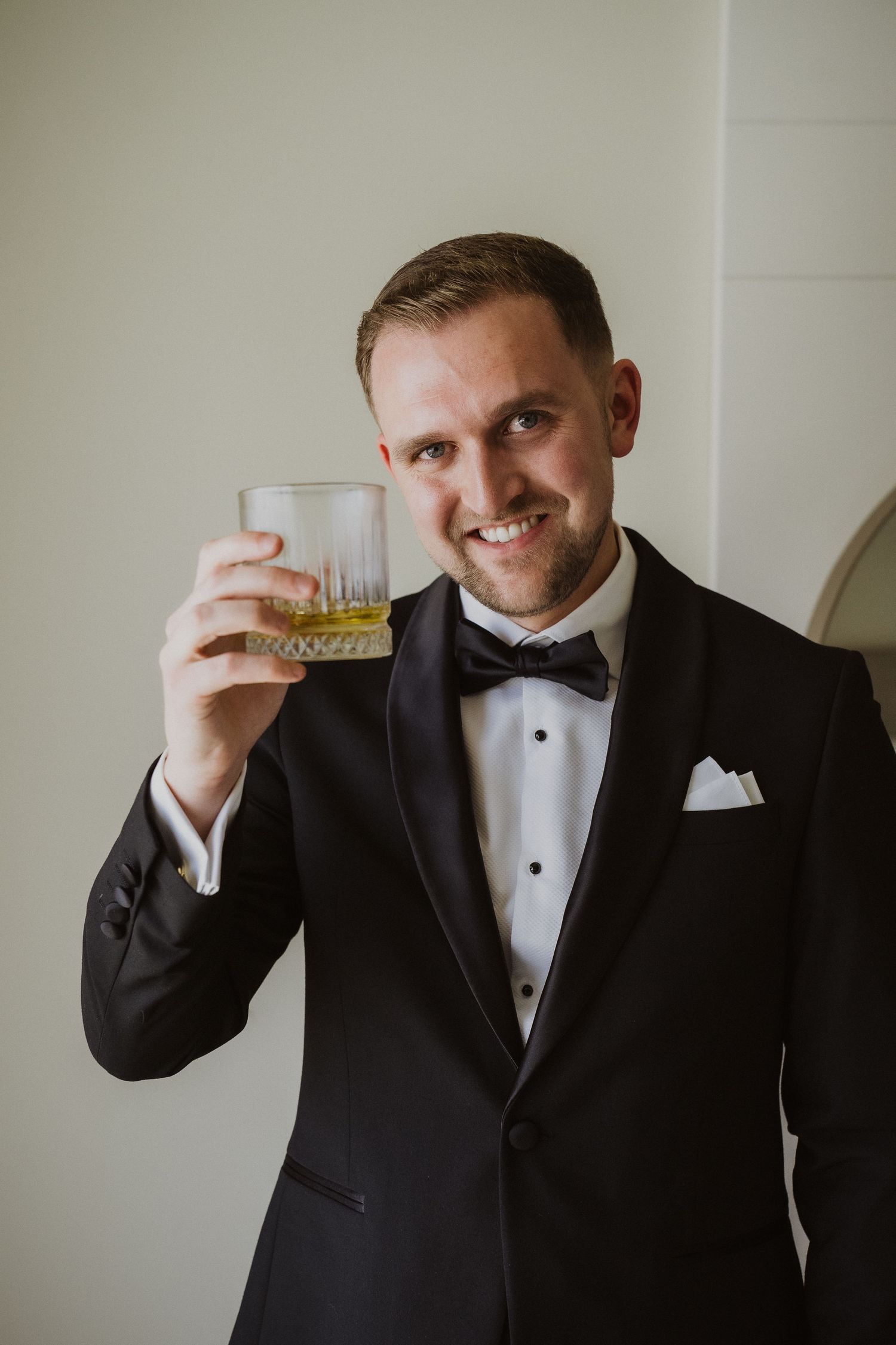 Smiling groom in black tuxedo raising glass of whiskey in celebratory toast before wedding.