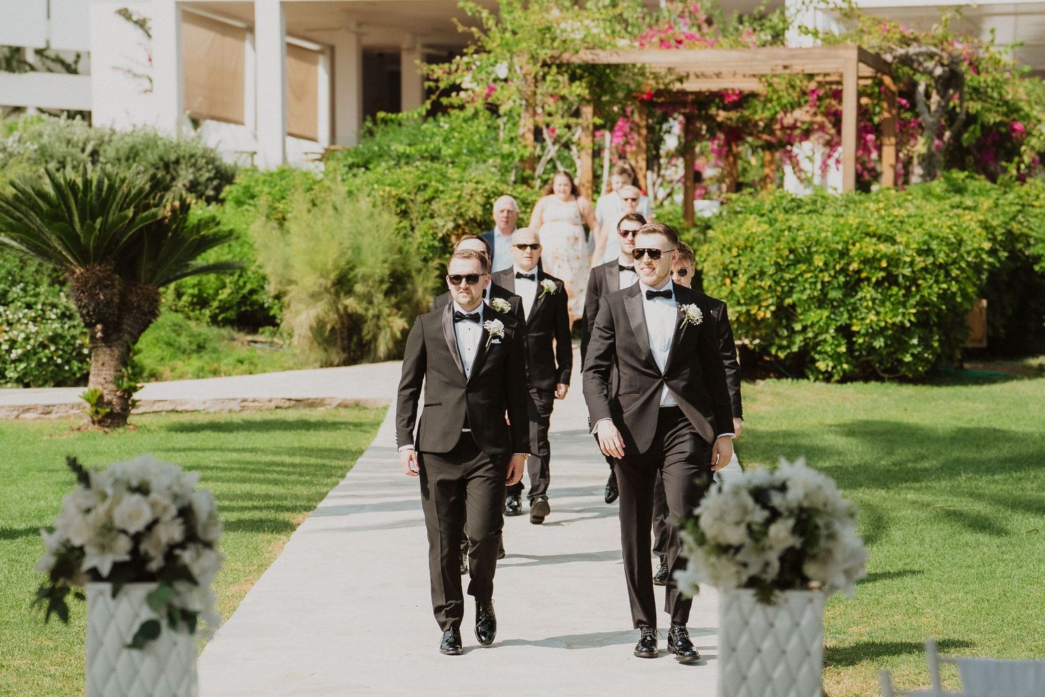 Groomsmen in black tuxedos and sunglasses walk down outdoor pathway at resort wedding venue.
