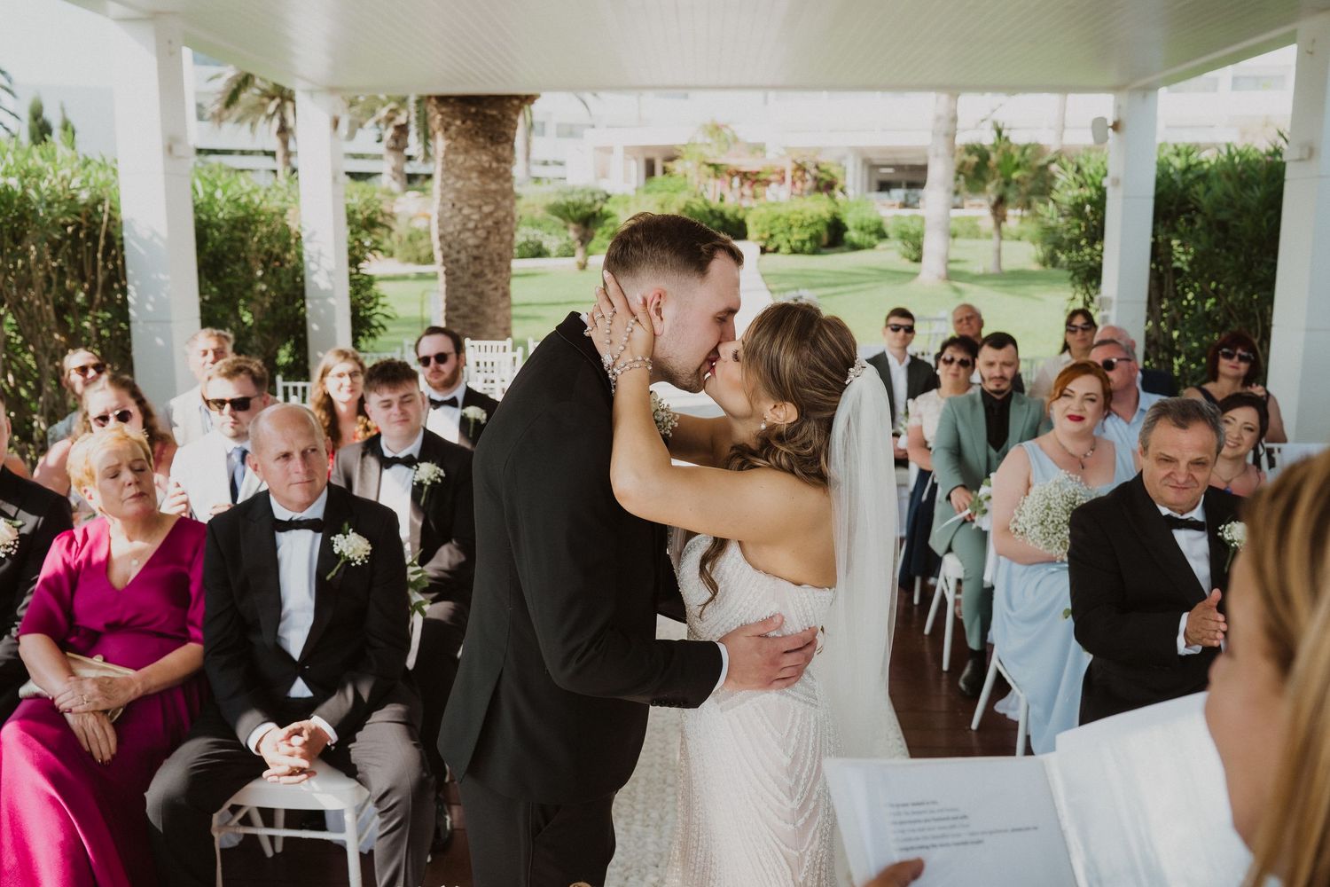 Bride and groom share first kiss at outdoor wedding ceremony with smiling guests.