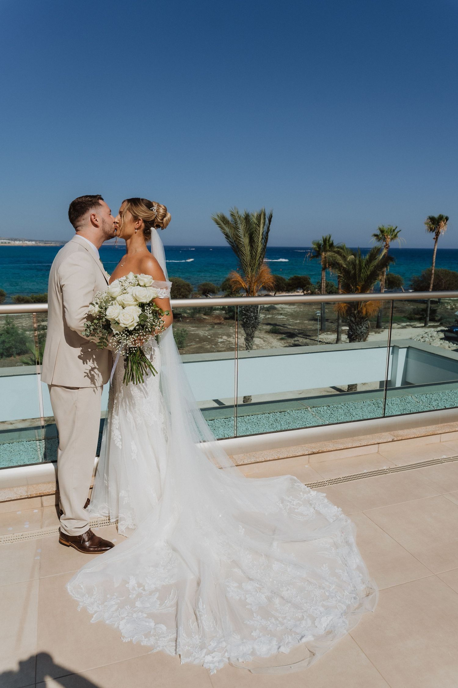 A bride and groom share a romantic moment on a tropical balcony overlooking turquoise waters and palm trees.
