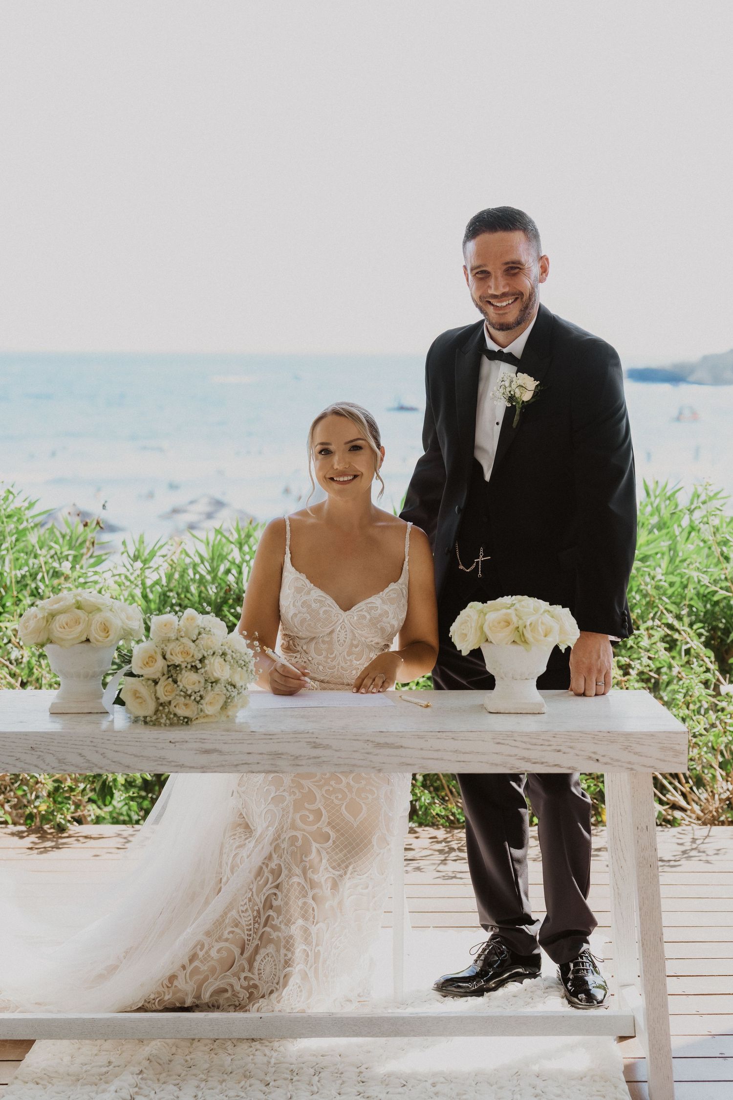 Newlyweds sign marriage documents at an oceanside ceremony table.