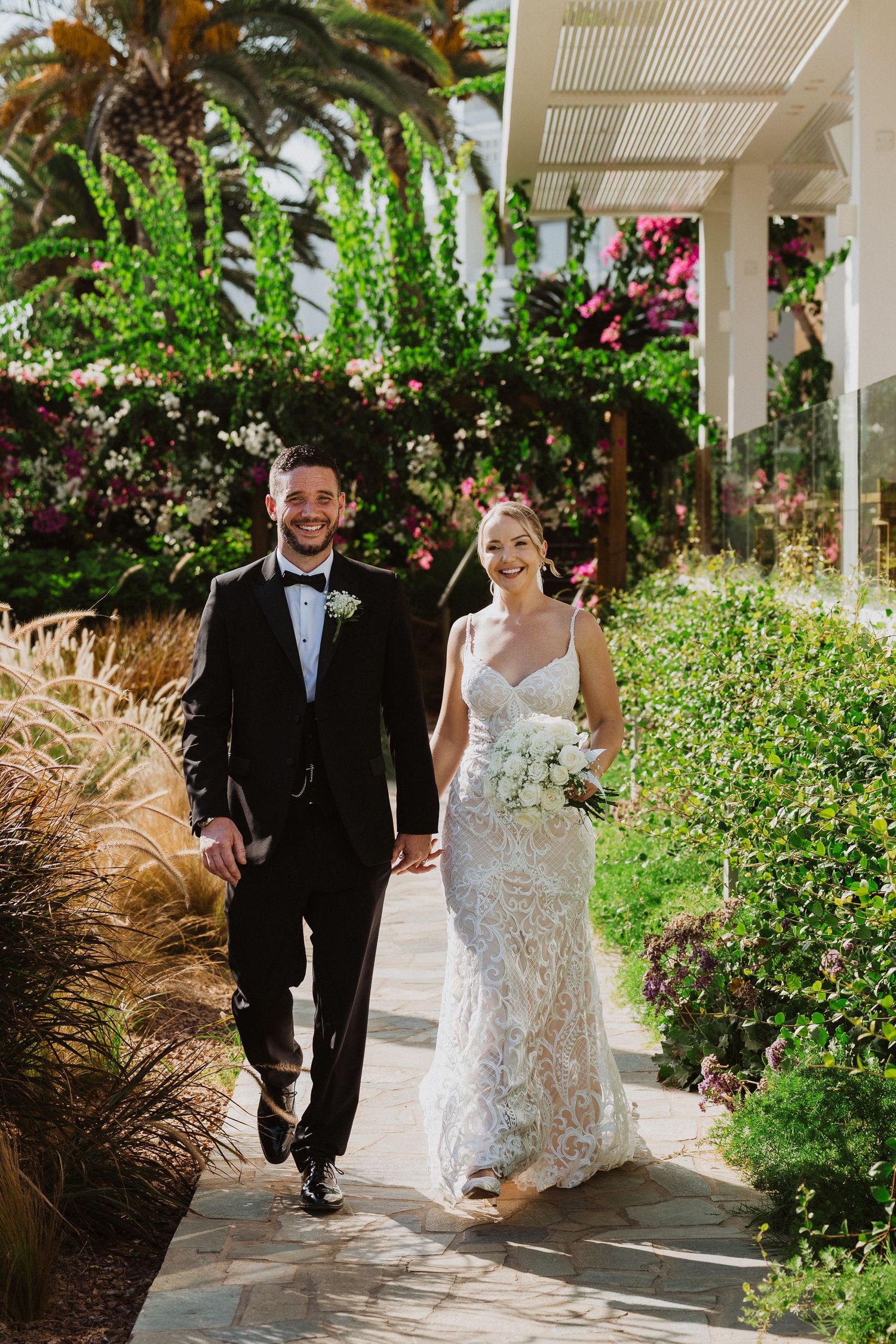 Newlyweds stroll down a tropical garden pathway lined with ornamental grasses and flowering plants.