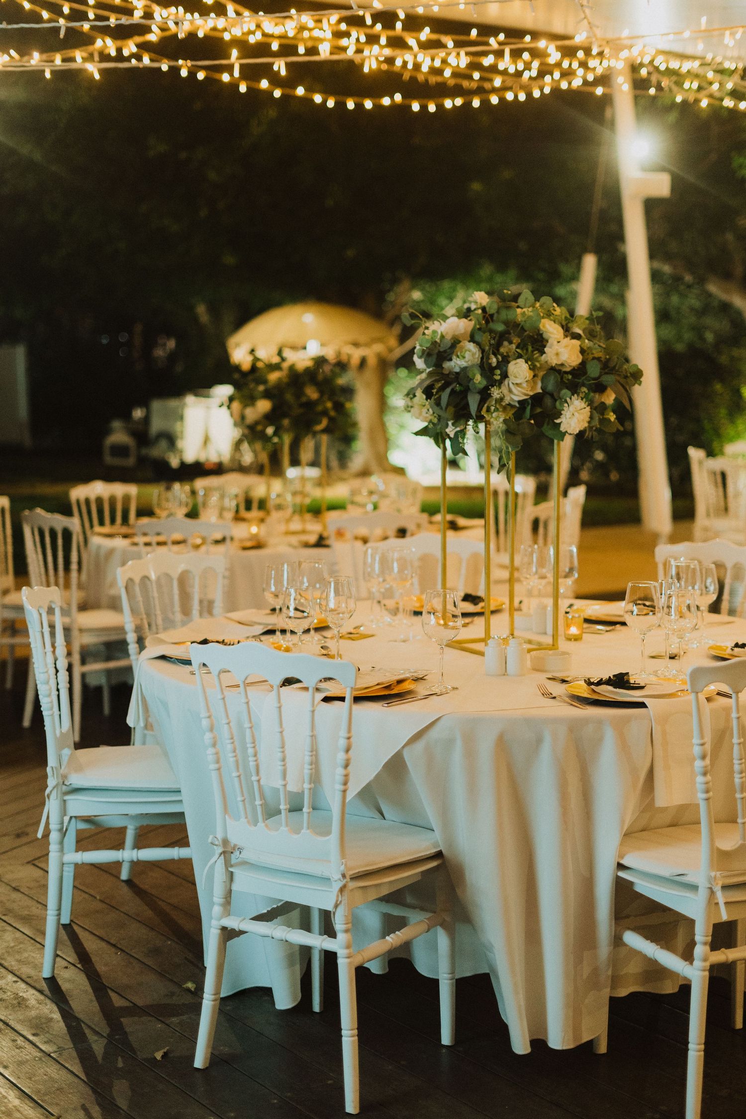 Wedding reception tables with white linens and tall floral arrangements illuminated by overhead string lights.