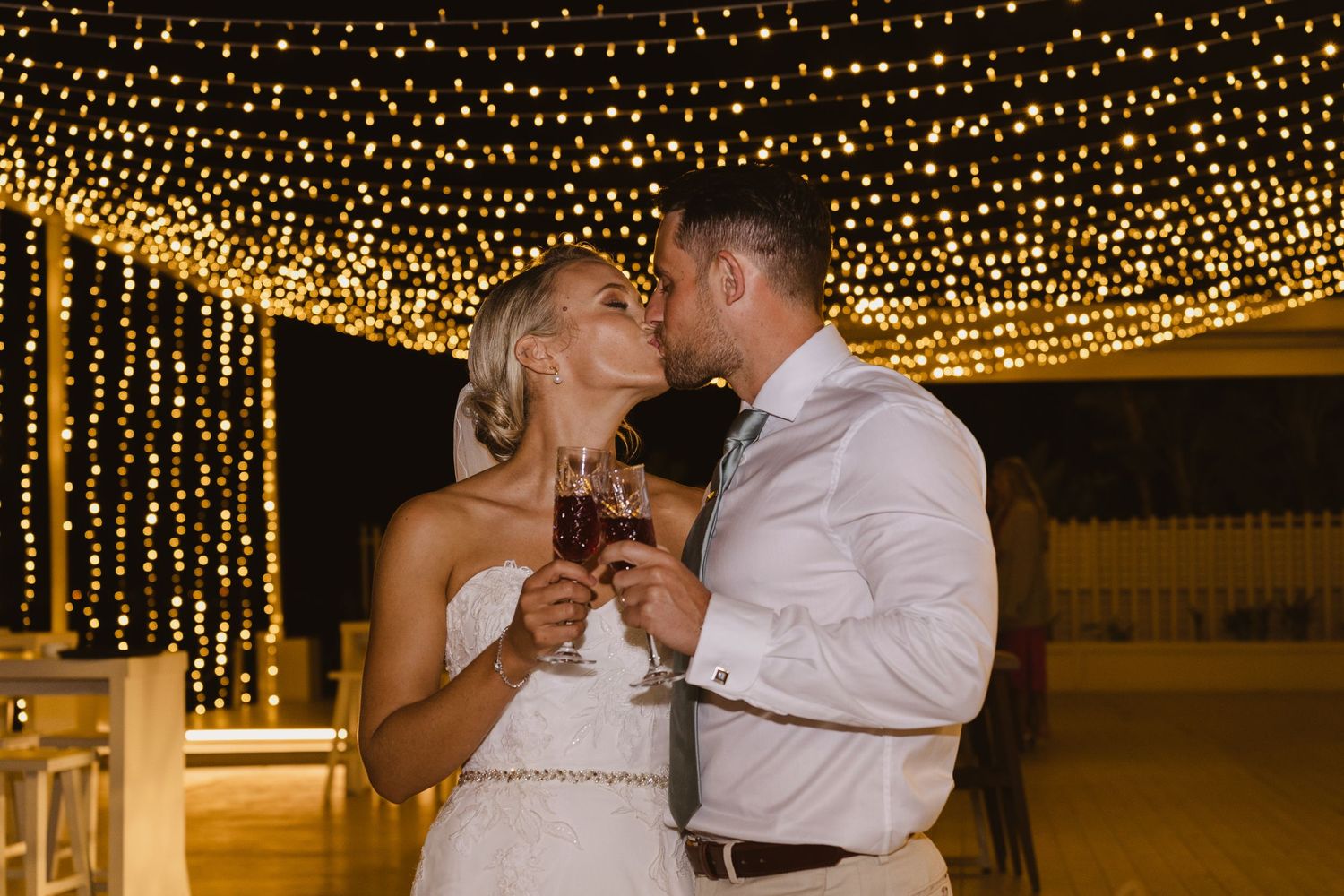 Romantic wedding kiss shared during champagne toast under canopy of golden lights.