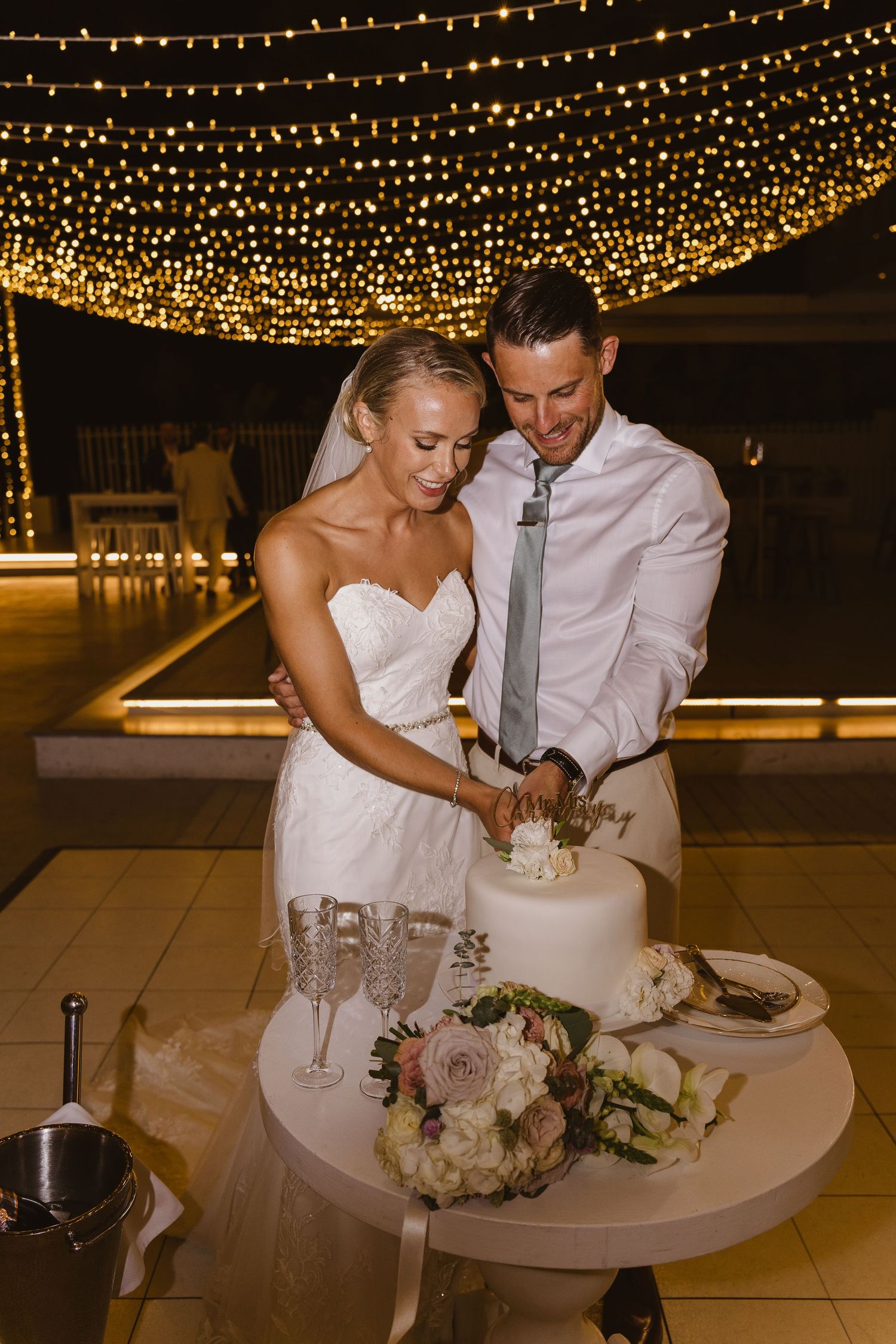 A couple shares a cake cutting moment under a magical ceiling of string lights.
