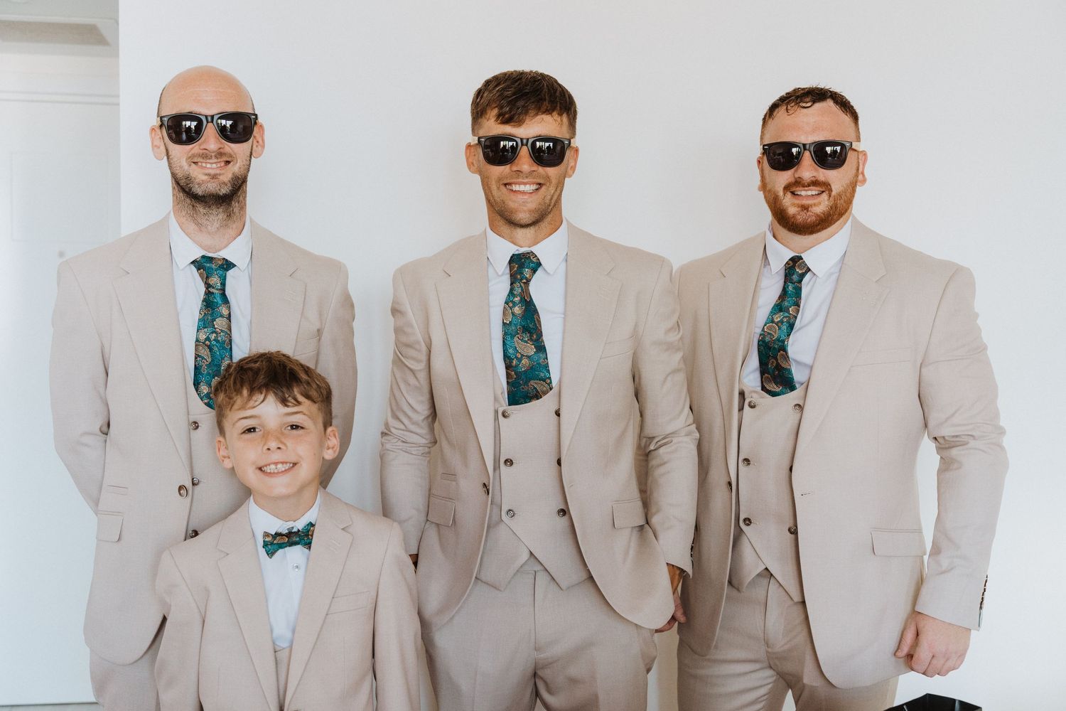 Group wearing matching beige suits and patterned ties posing for formal photo at wedding.