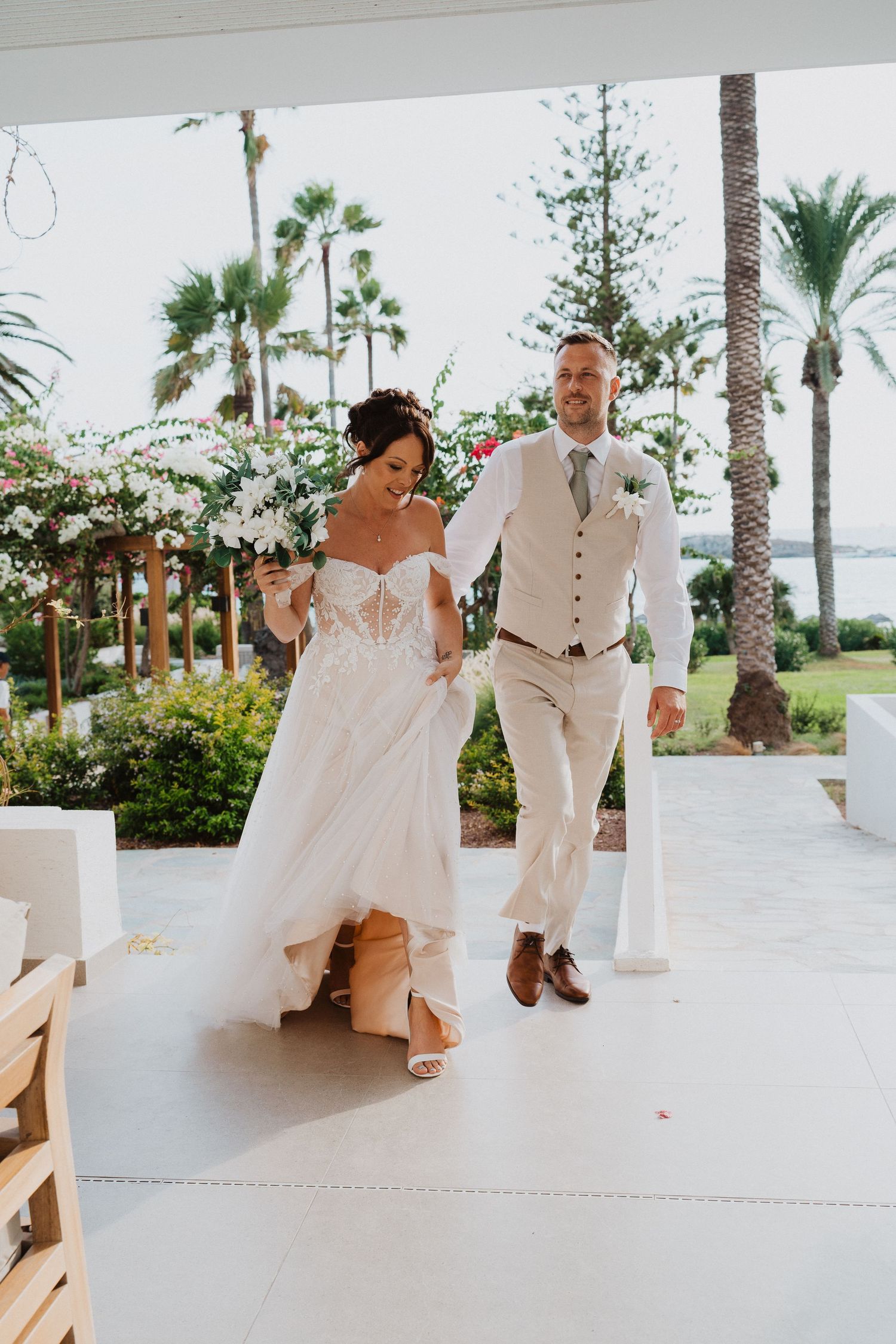 A couple walks together at a tropical wedding venue with palm trees and white floral decorations along a covered walkway.