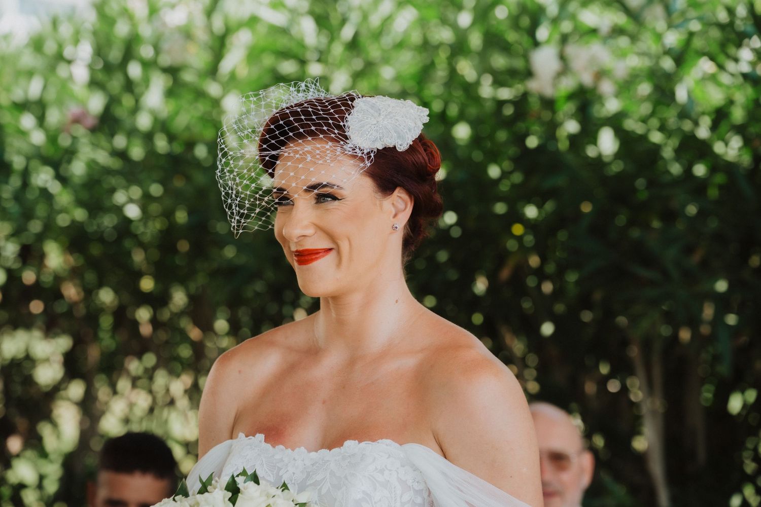 A bride wearing a vintage-inspired birdcage veil smiles during an outdoor wedding celebration.
