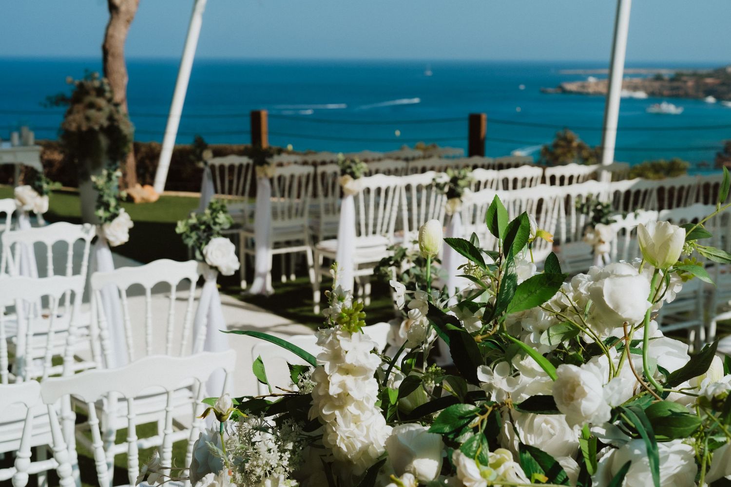 White floral centerpieces and chairs arranged at a coastal wedding venue with stunning turquoise ocean views.