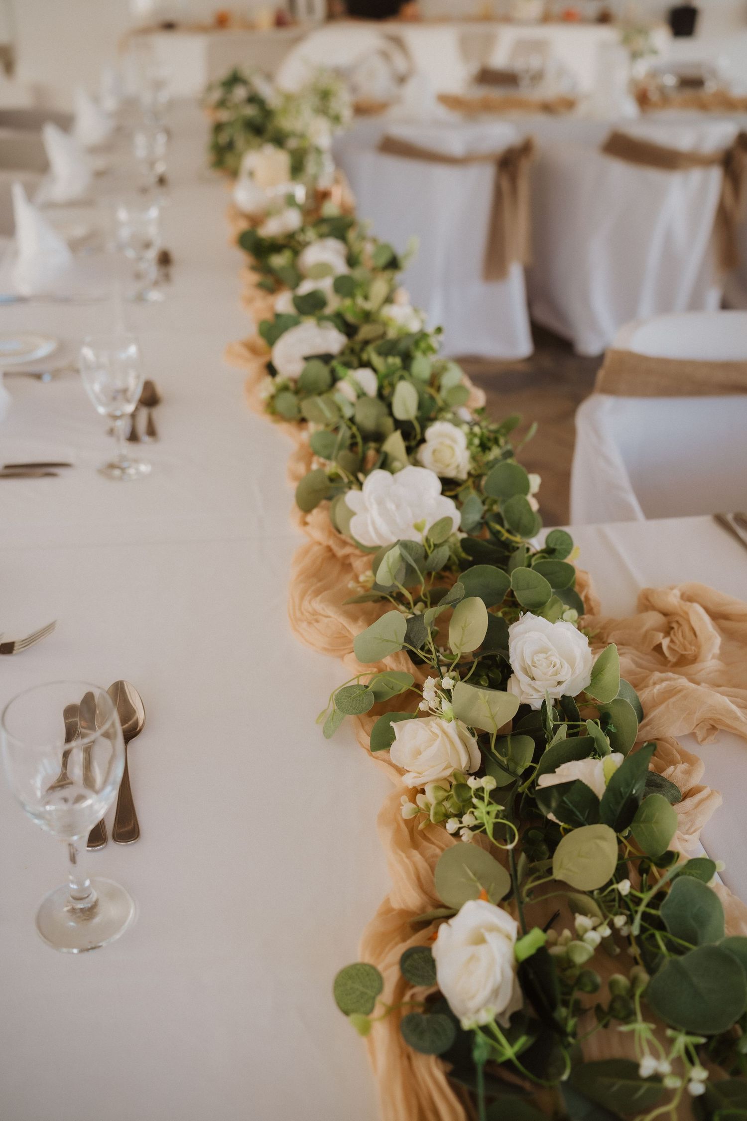 White roses and greenery line a wedding table runner with elegant place settings and glassware.