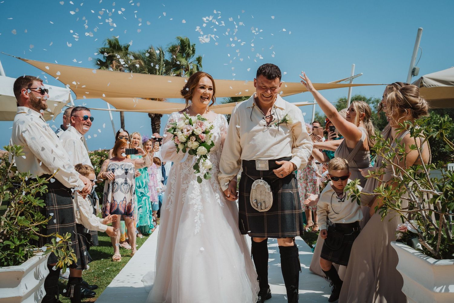Scottish wedding celebration with traditional kilts takes place outdoors under beige canopy with palm trees and ocean view.