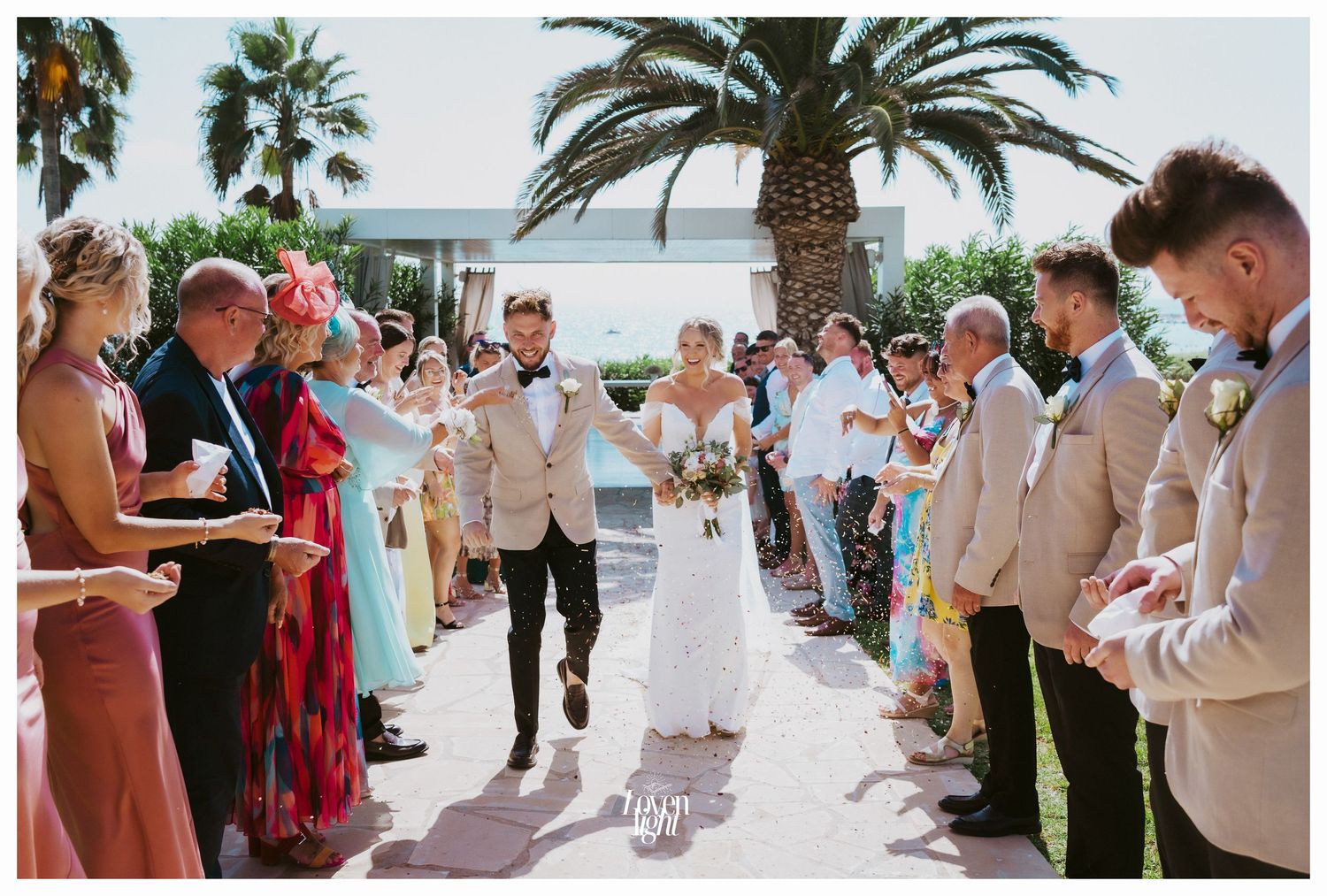 Wedding party lines an outdoor walkway with palm trees while celebrating the newlyweds.