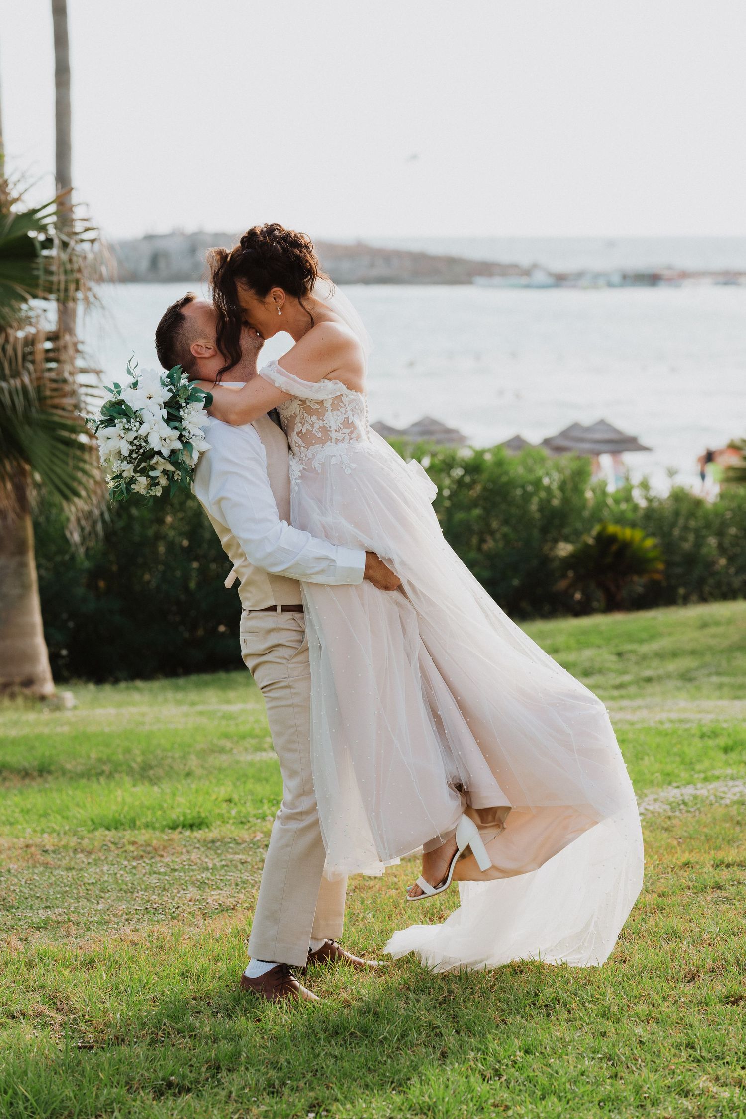 A wedding couple shares a romantic embrace on a grassy hillside overlooking the ocean at their tropical destination ceremony.