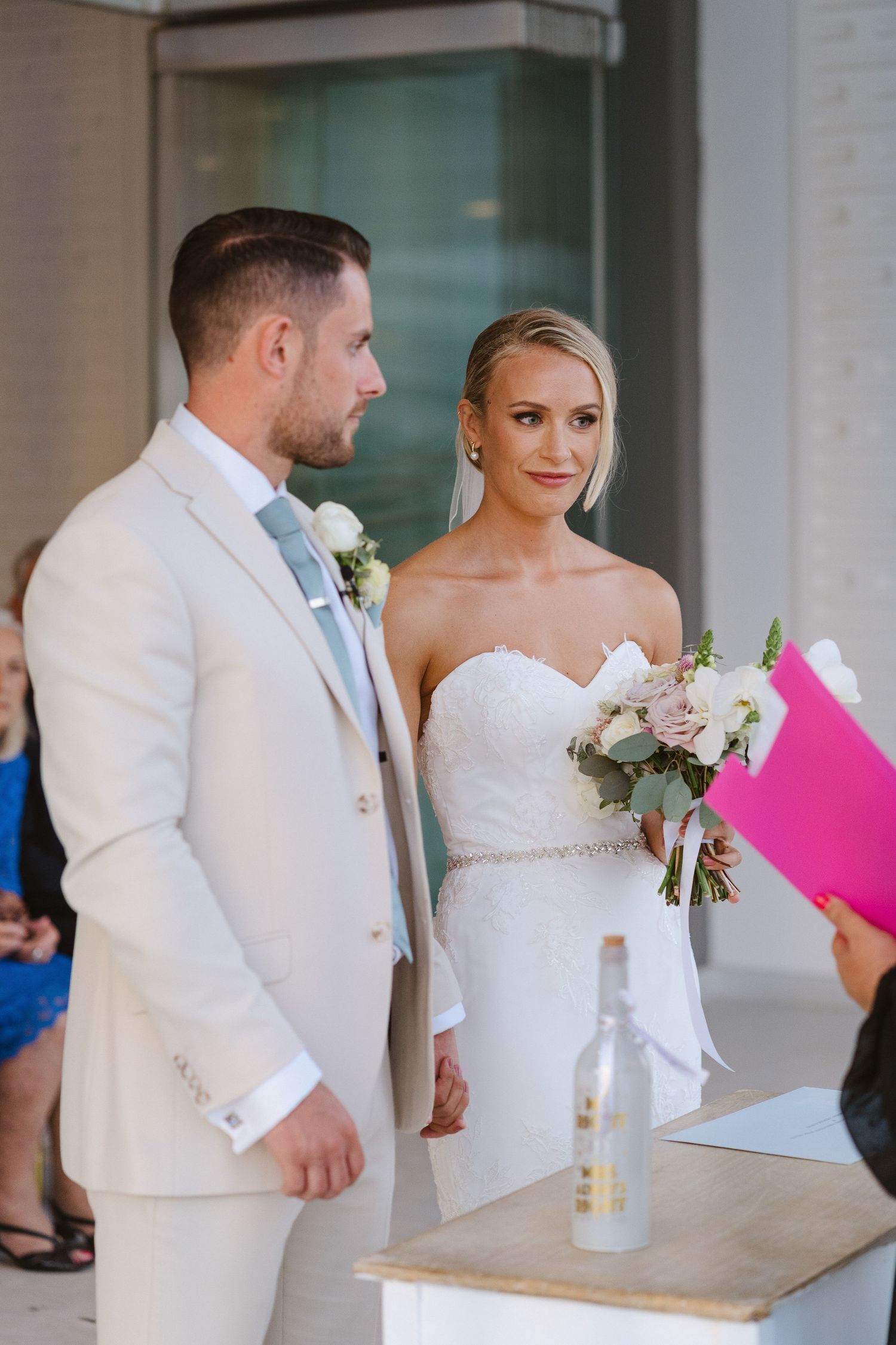 Couple in white attire standing together during wedding ceremony with delicate floral bouquet.