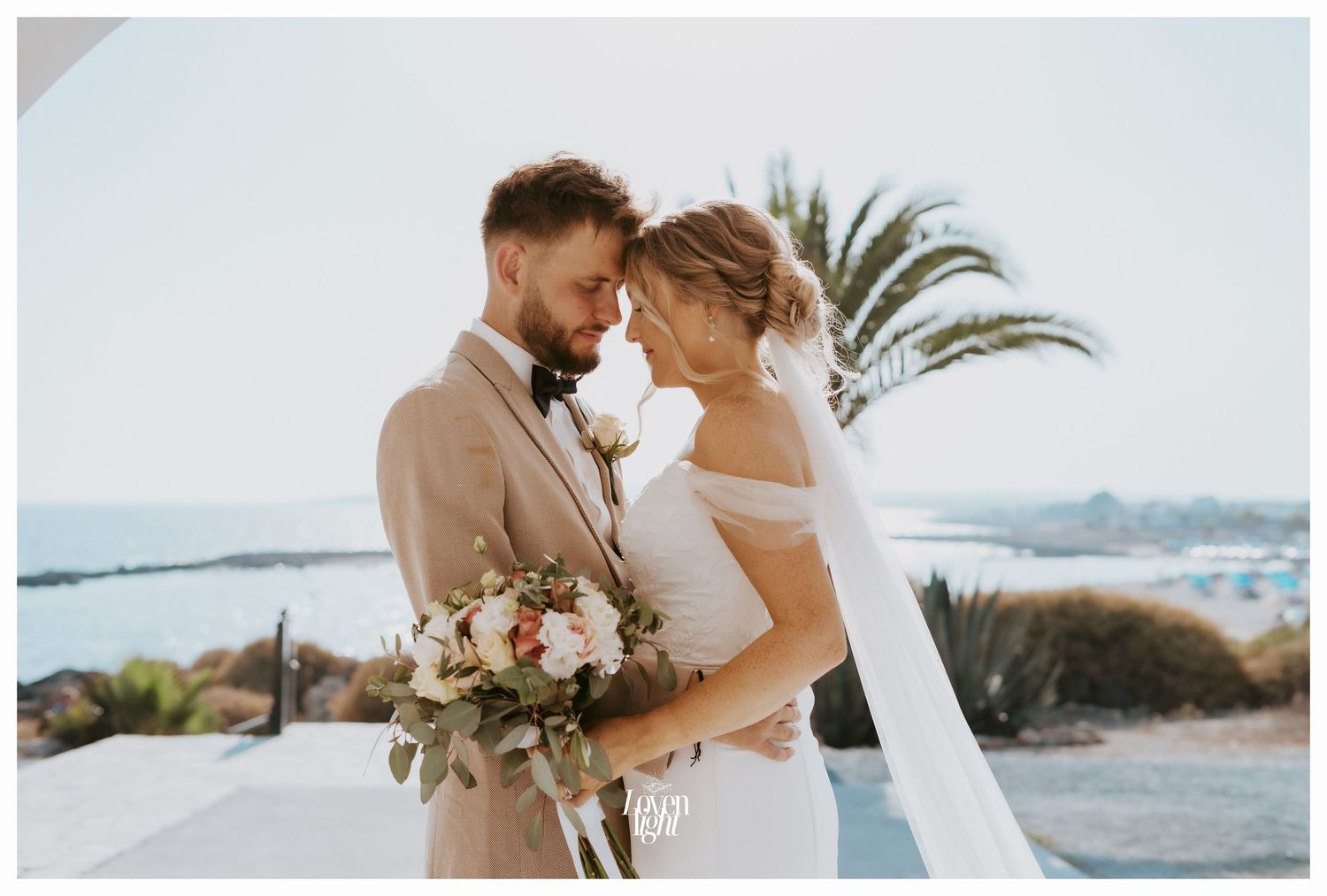 Newlyweds share intimate moment at coastal wedding venue with palm trees and ocean views.