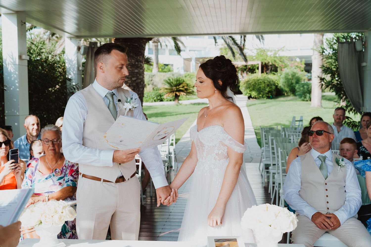 A couple shares a touching moment during their outdoor wedding ceremony under a covered porch on a sunny day.