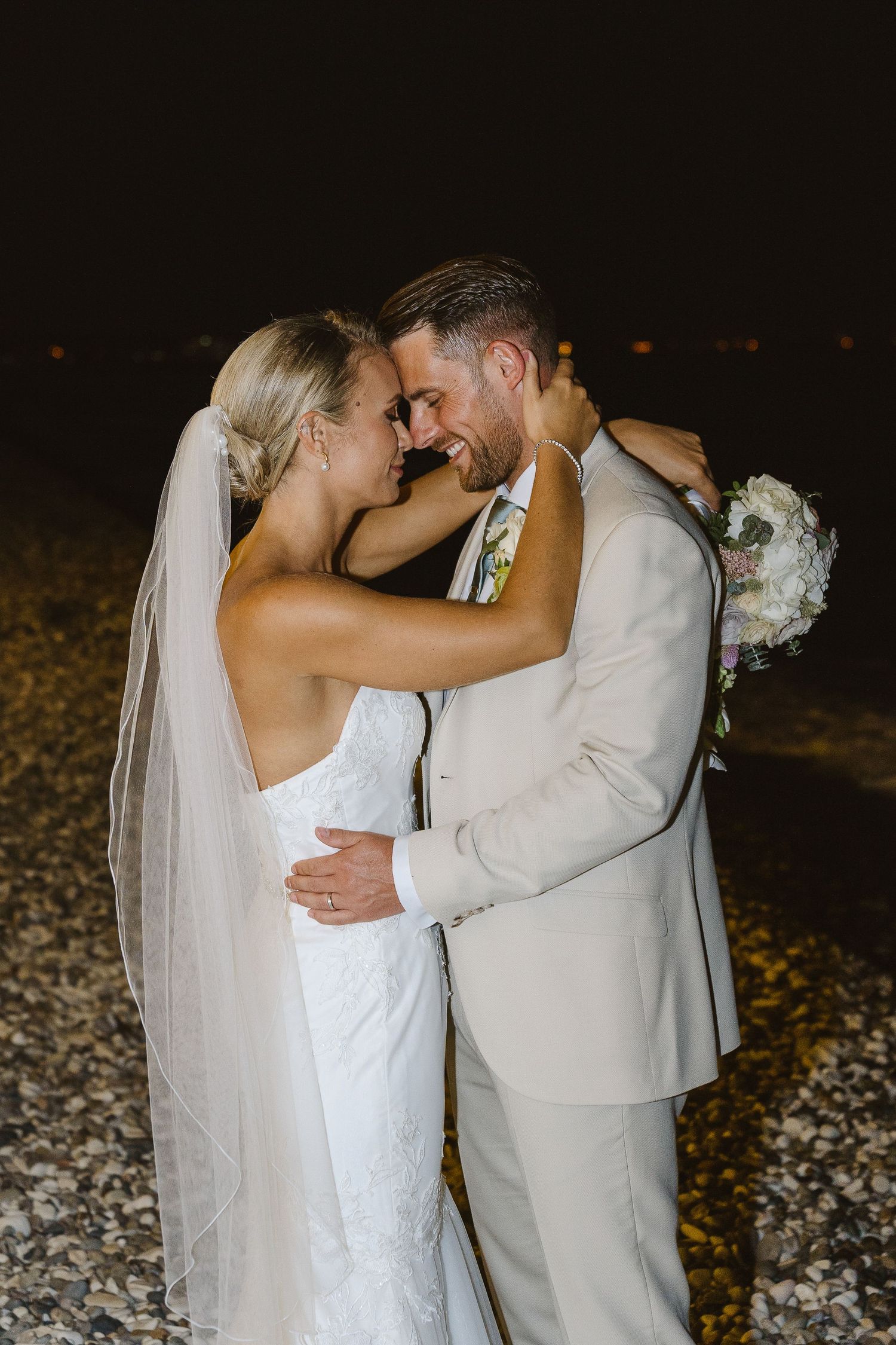 Romantic wedding couple sharing an intimate moment on a beach at night with bride holding white bouquet.