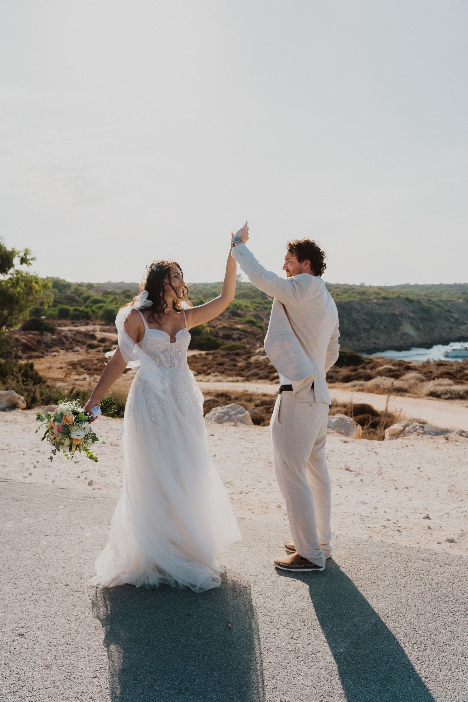 A couple in white wedding attire dance together on a scenic coastal cliff overlooking the ocean at sunset.