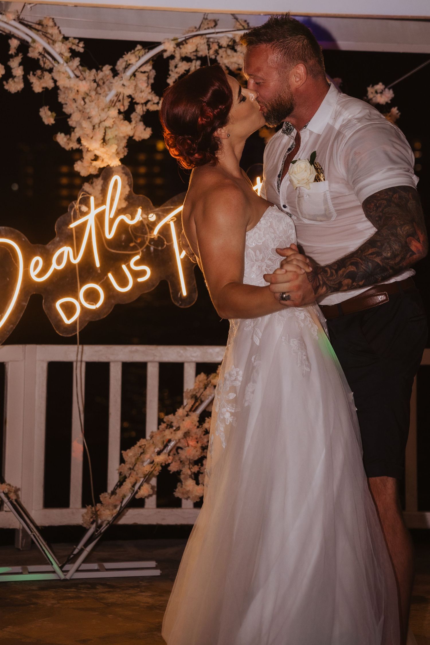 Newlyweds share romantic dance under neon 'Death do us part' sign with white floral decorations.