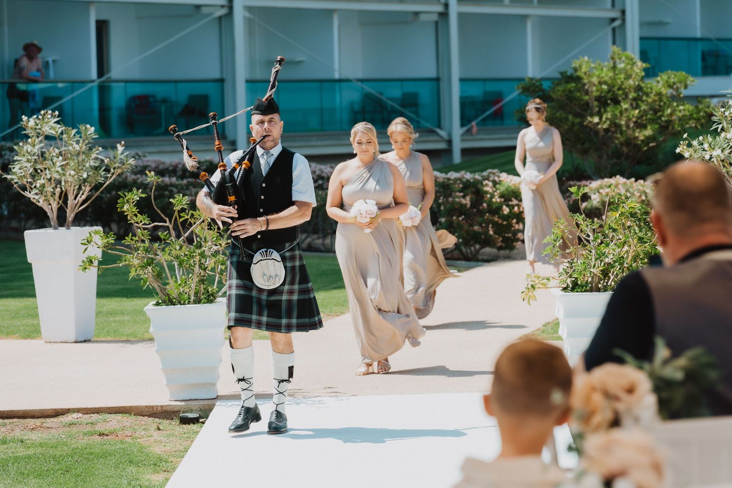 Bagpiper leads wedding processional down outdoor aisle with decorative planters.