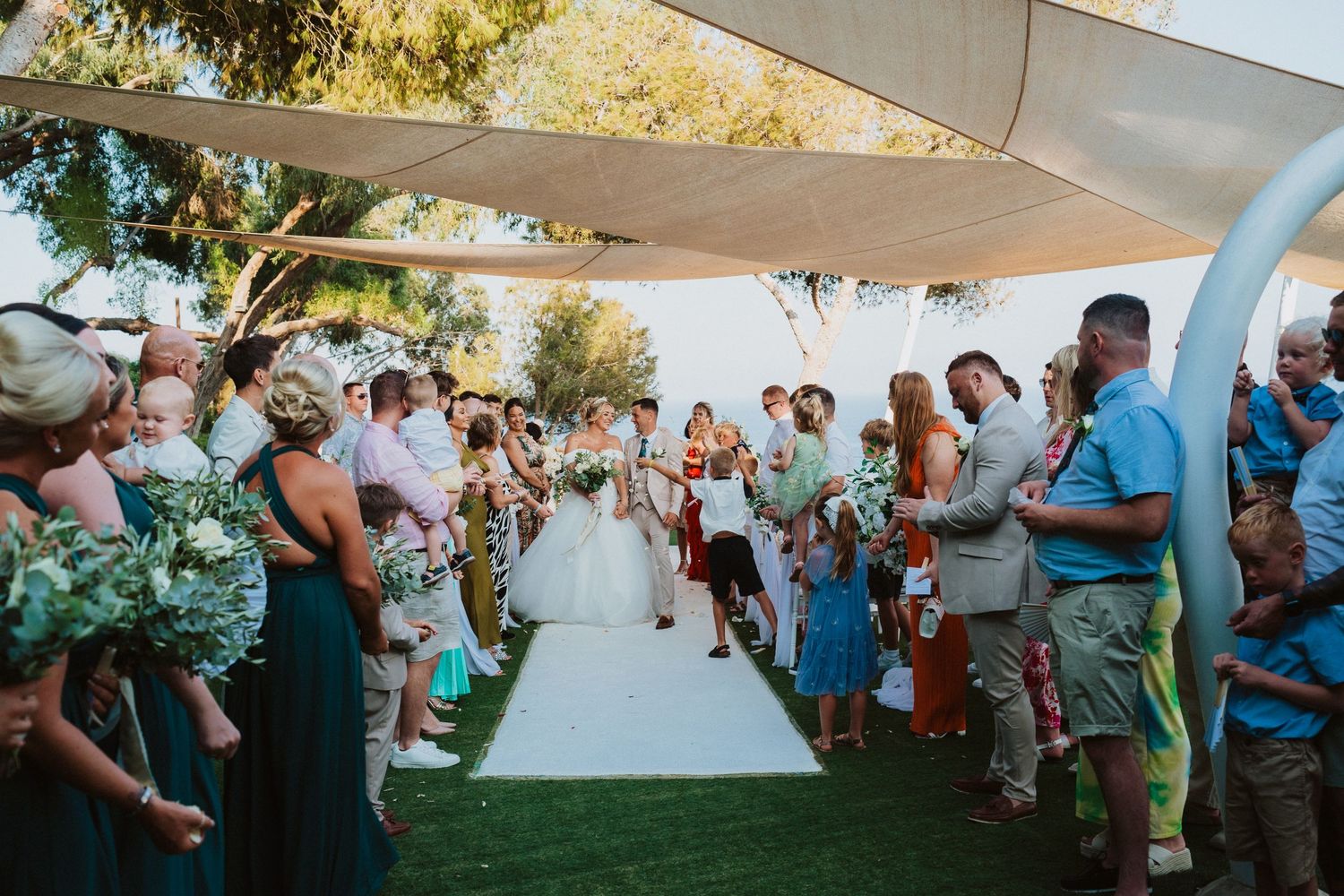Wedding guests line an outdoor aisle under white canopies during a ceremony.