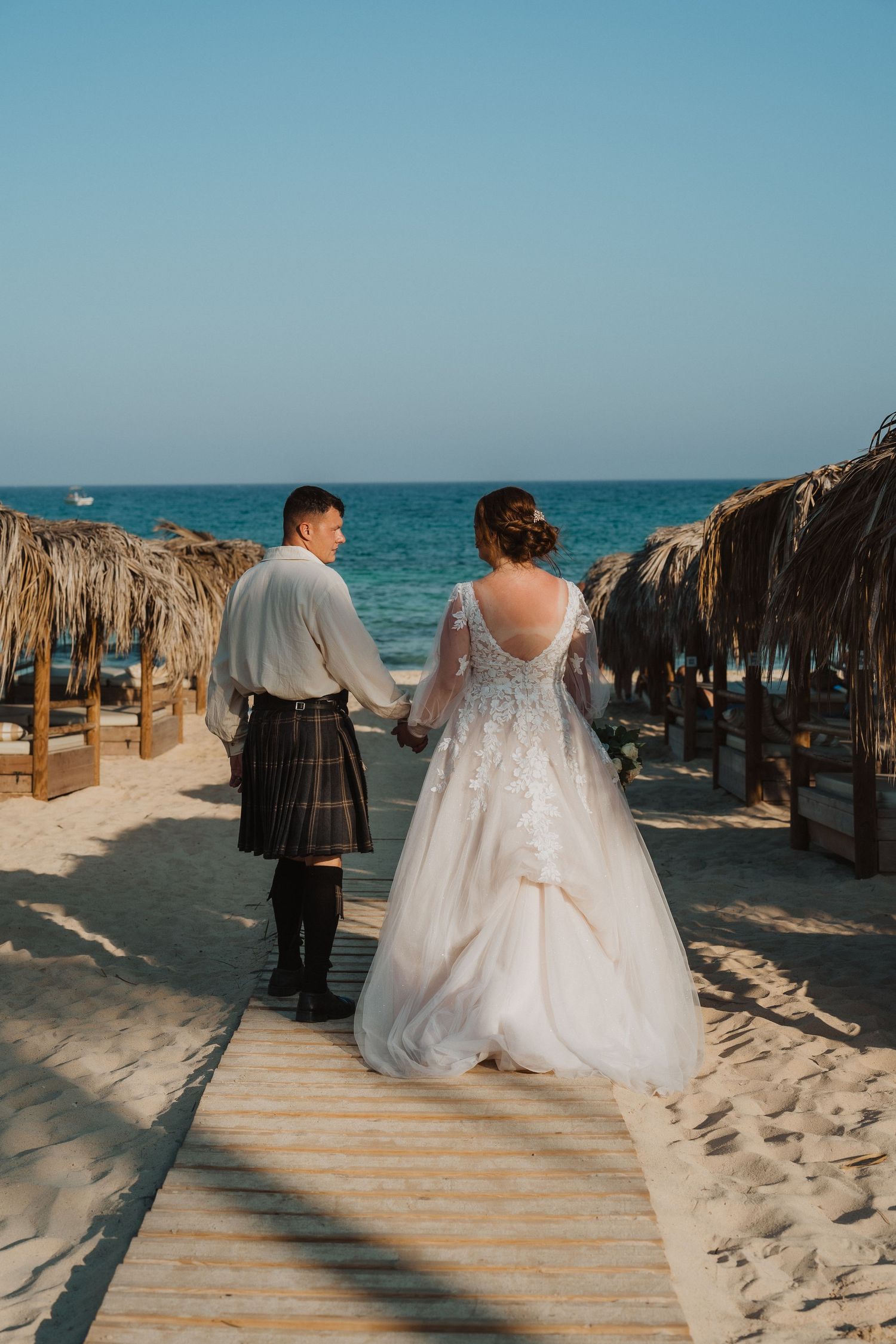 A wedding couple walks along a wooden boardwalk between beach umbrellas by the ocean.