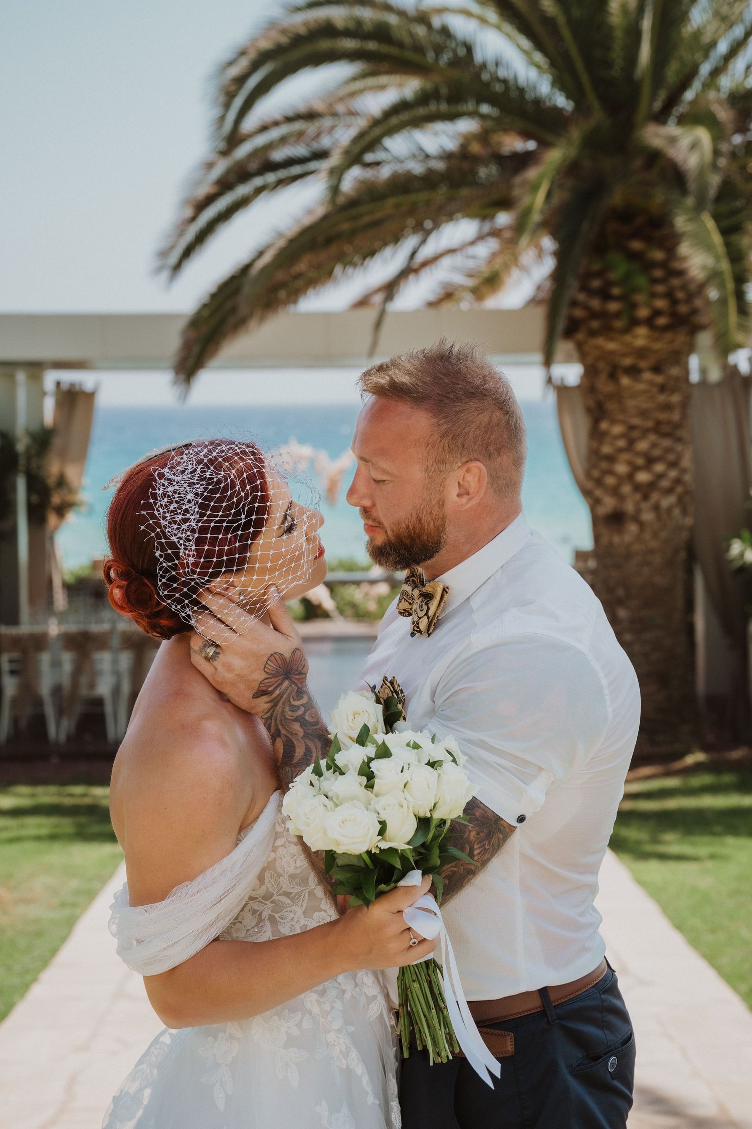 Tender moment between newlyweds holding white bouquet with beach wedding setup in background.