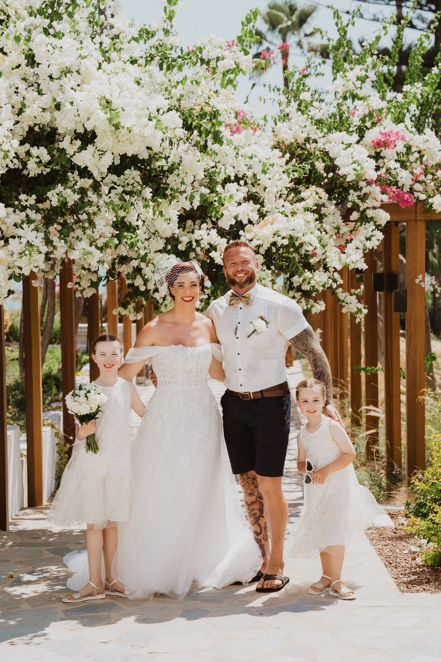 Wedding group poses under white flowering pergola against tropical backdrop.