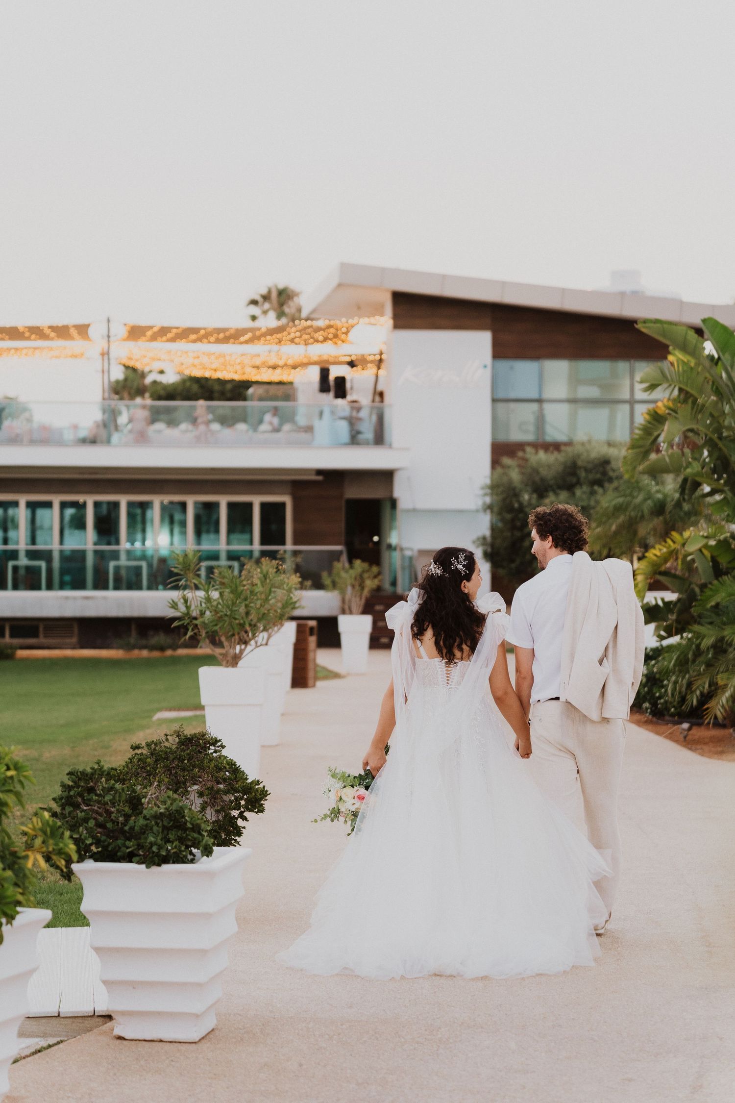 A couple walks along a modern resort pathway with white planters and lush greenery at sunset.