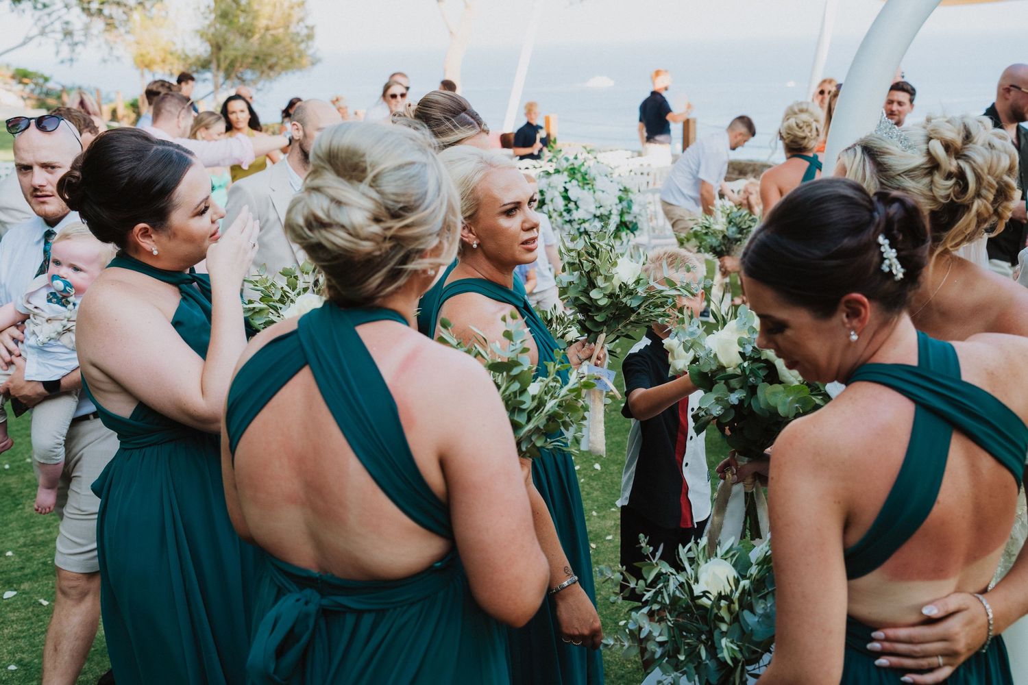 Wedding guests in emerald green dresses gather outdoors holding bouquets of fresh greenery at a celebration.