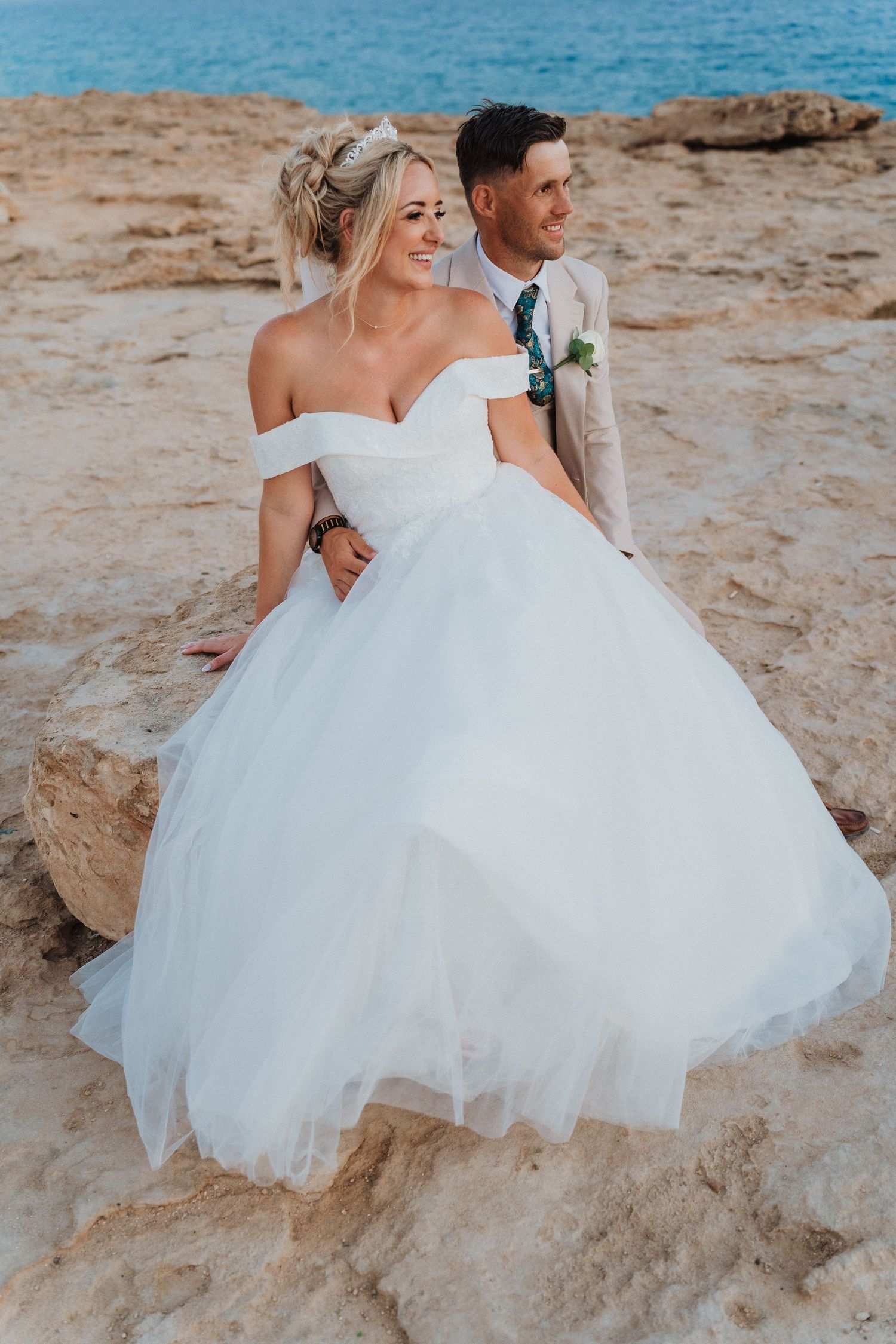A bride in an off-shoulder white ball gown and groom standing together on a sandy beach during a coastal wedding.