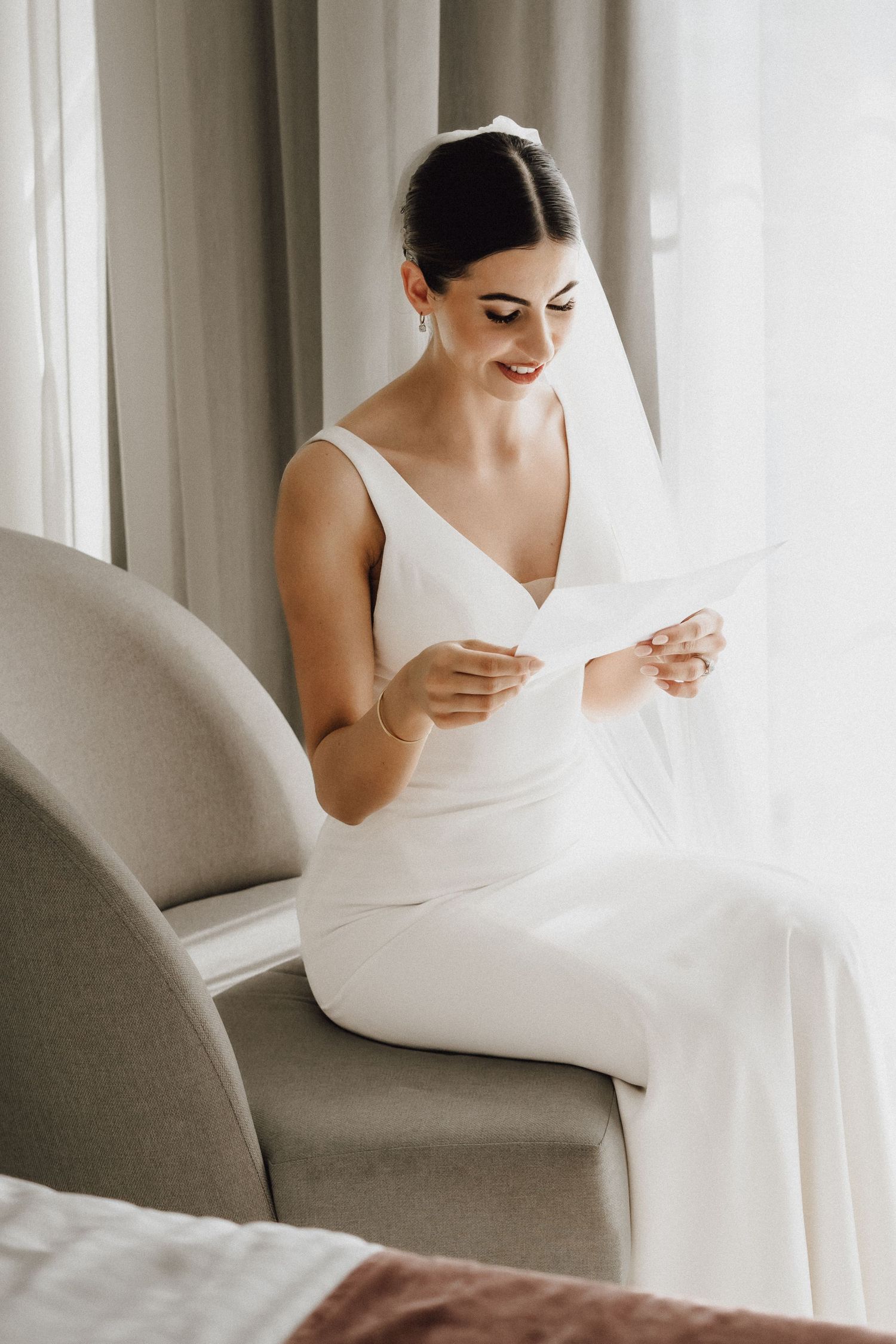 Bride in elegant white dress sits reading a note in a modern room with natural light.