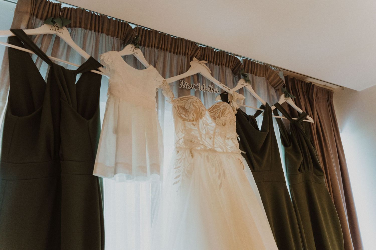 Wedding attire including a white bridal gown and dark bridesmaid dresses hanging on a curtain rod.