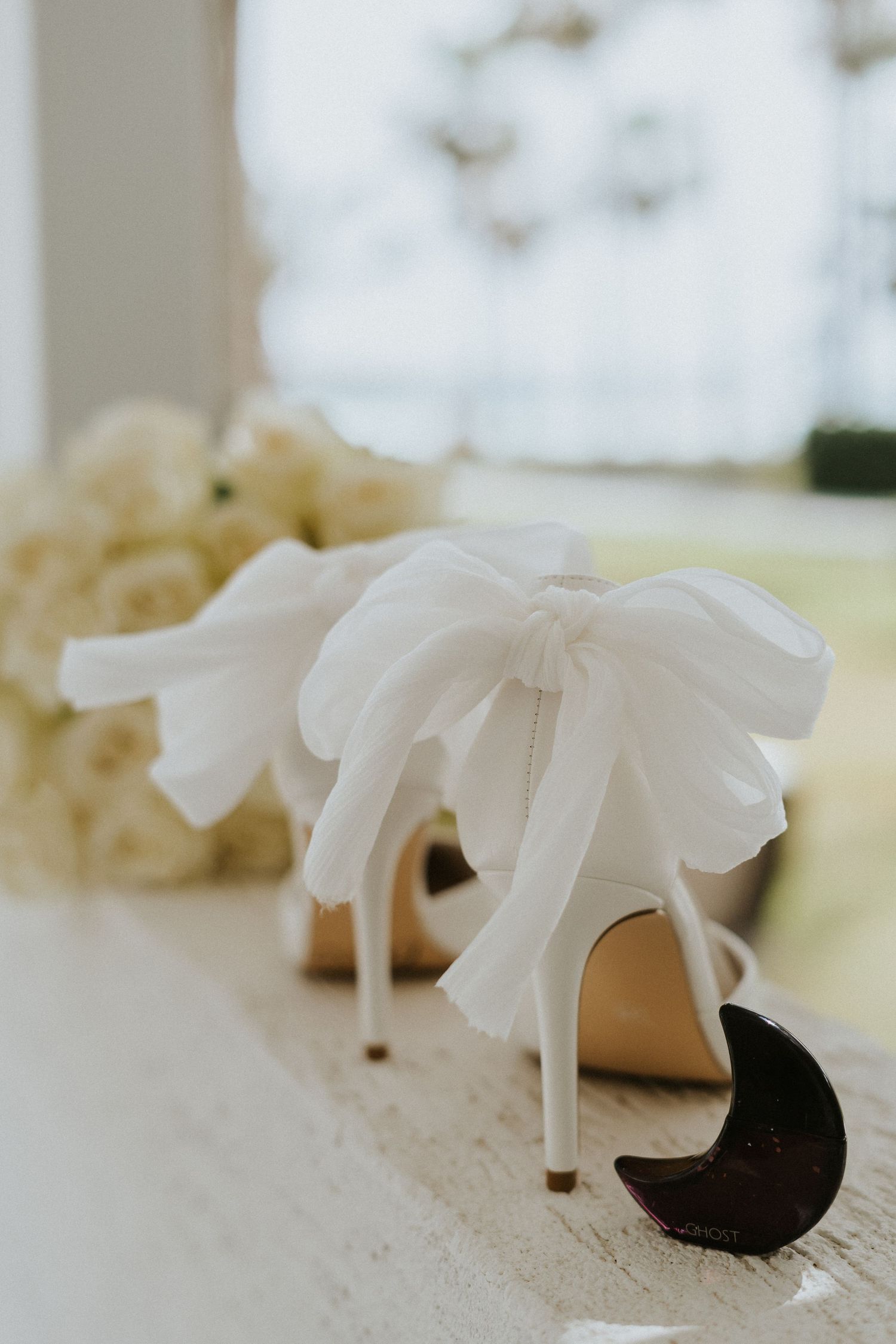 White bridal heels with bow detail photographed with soft roses in background.