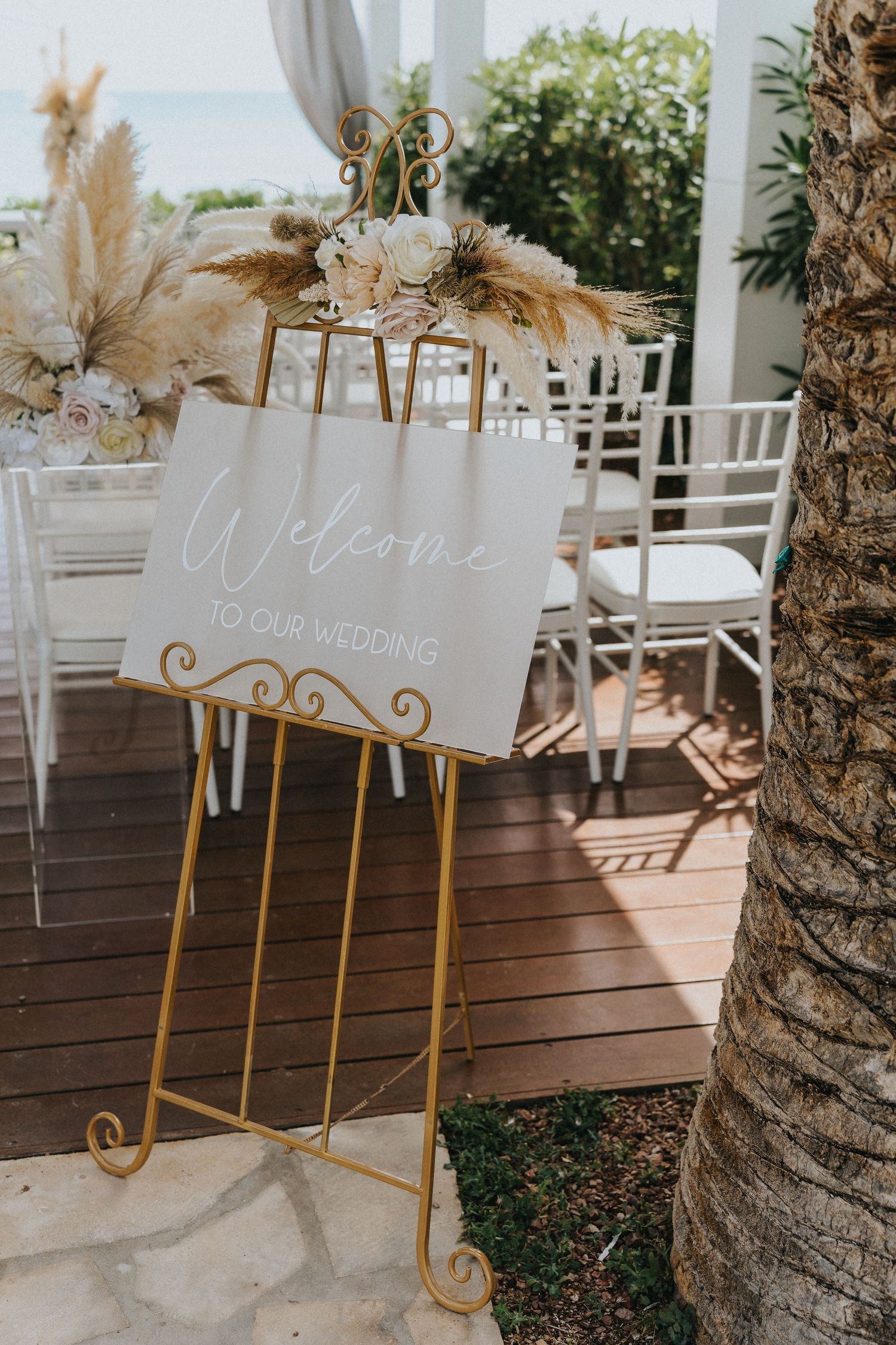 Elegant wedding welcome sign with gold accents and dried pampas grass decorations on a wooden deck by the ocean.