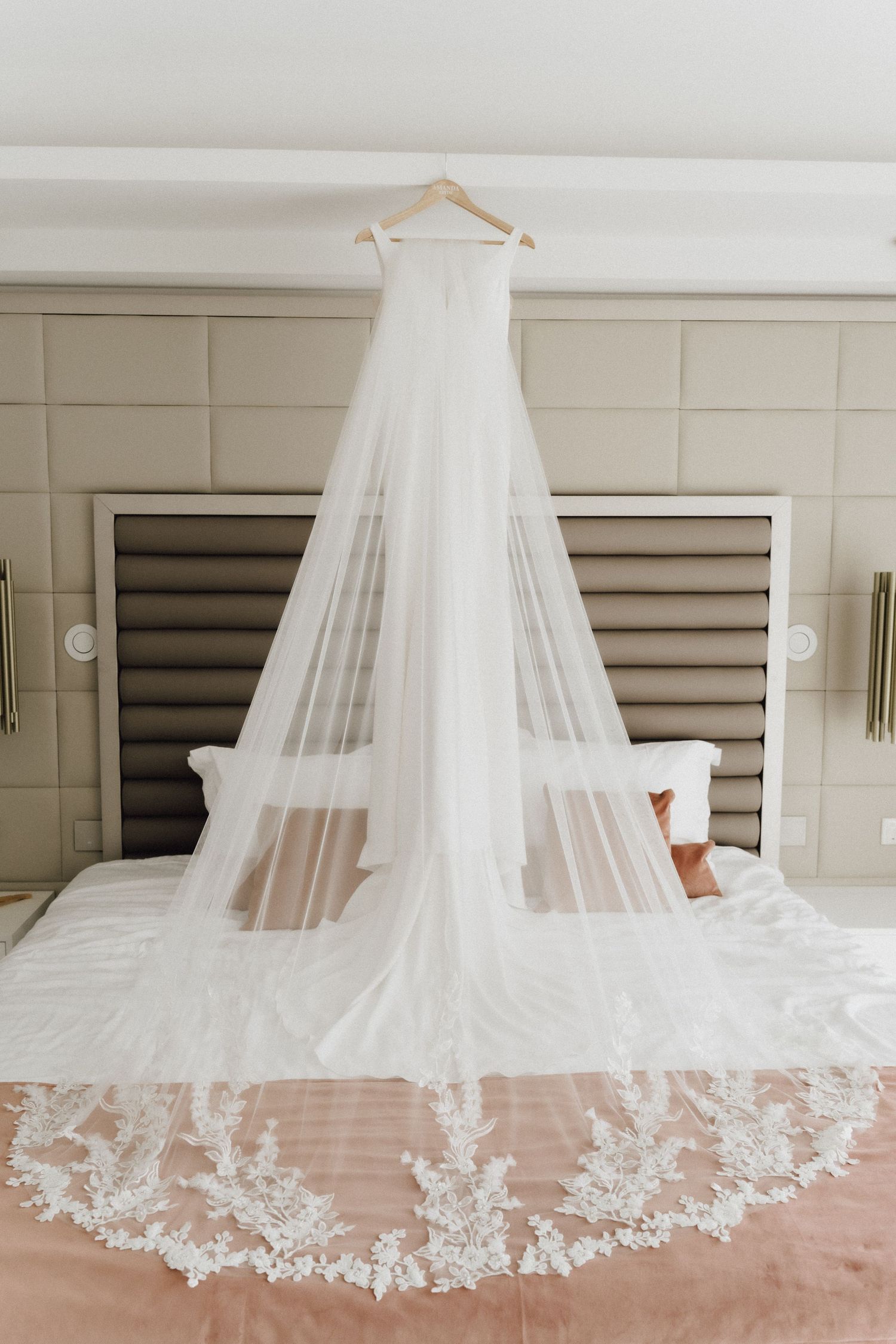 An elegant white wedding dress and veil with lace details hanging above a bed.