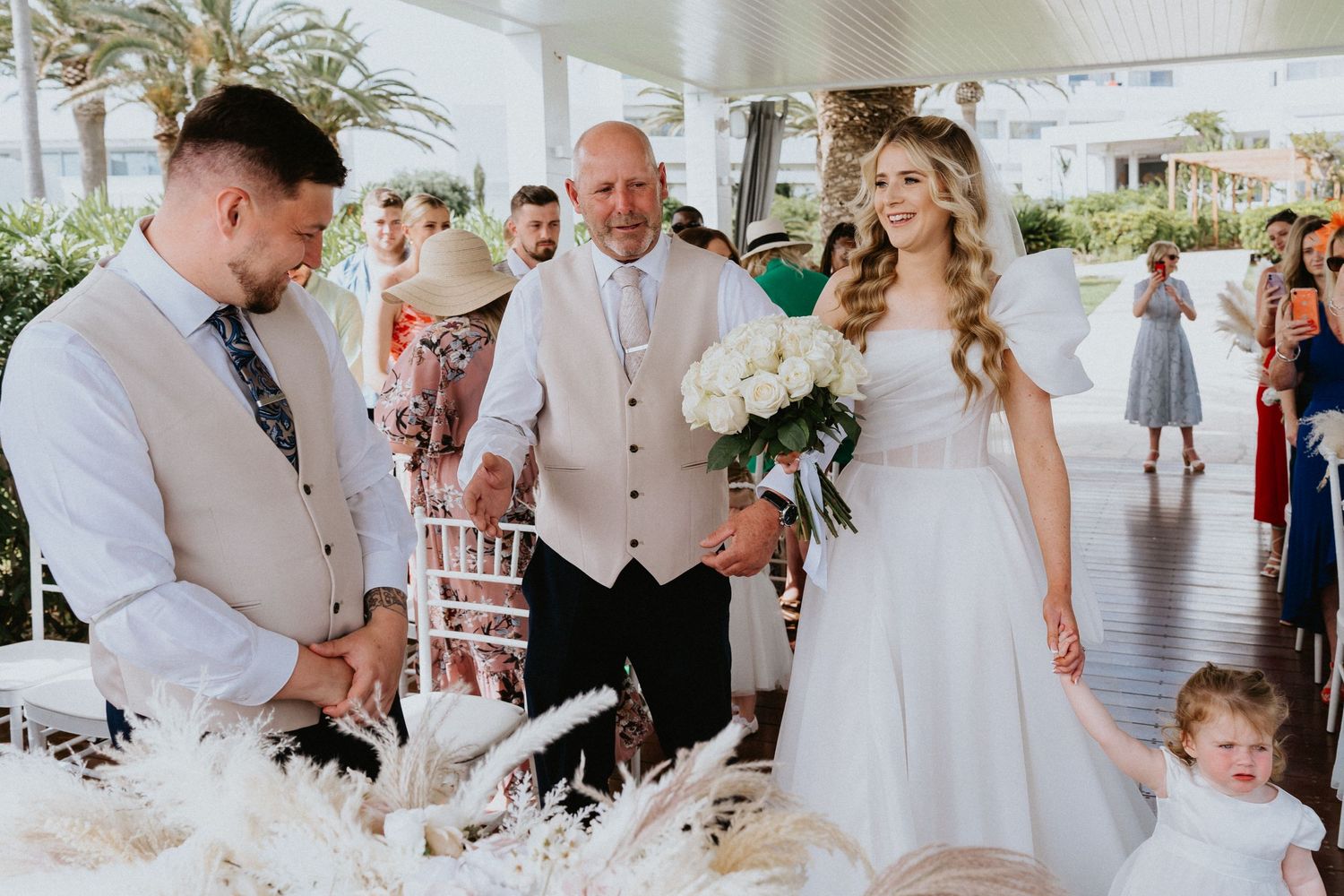Intimate outdoor wedding ceremony with white floral arrangements and tropical palm backdrop.