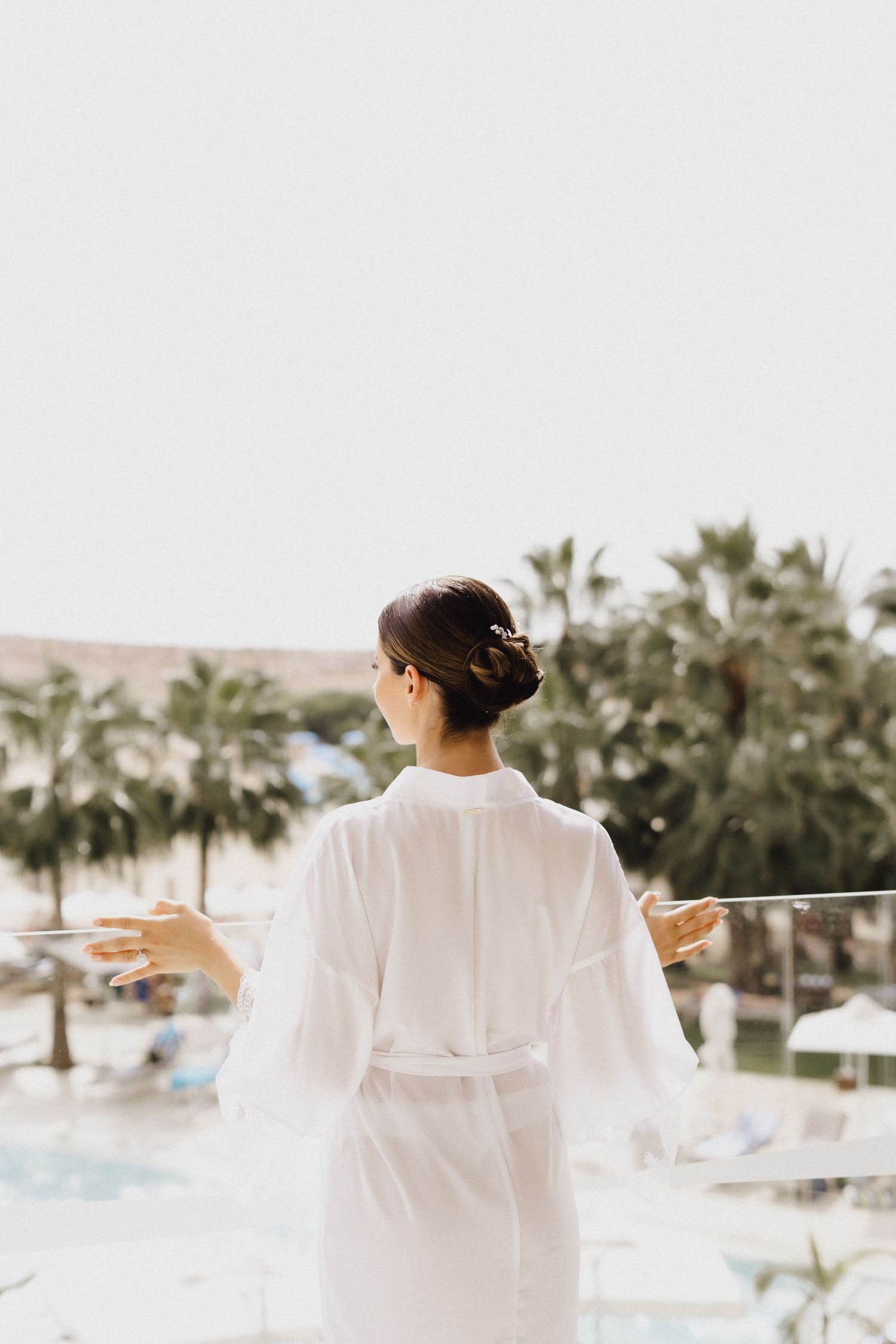 Back view of person in white robe gazing at tropical resort landscape from balcony.
