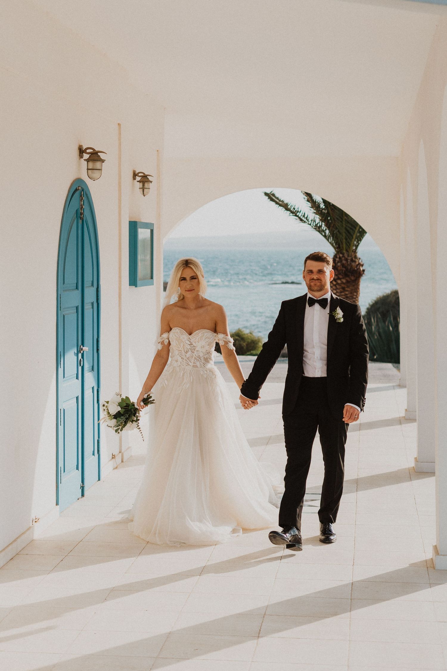 Newlyweds walk through Mediterranean archway with blue accents and ocean views.