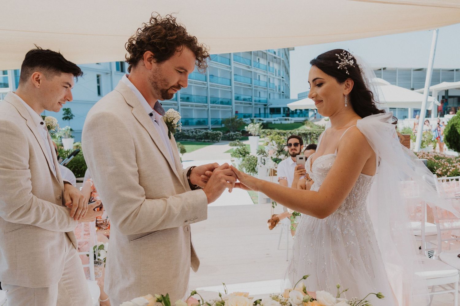 Intimate ring exchange moment during an outdoor wedding ceremony with beige and floral decor.