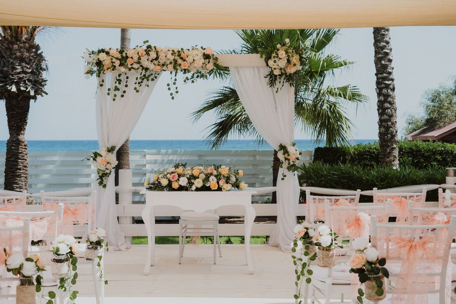 Beachfront wedding ceremony setup with white arch, peach-colored chairs, and tropical floral arrangements by the ocean.