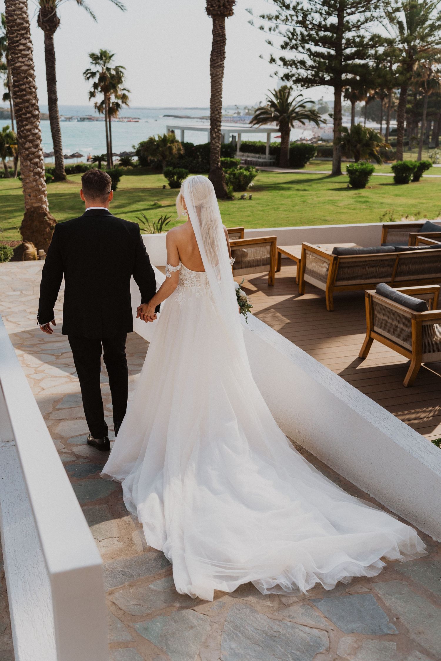 Elegant wedding couple walking on a seaside terrace with palm trees and ocean views.