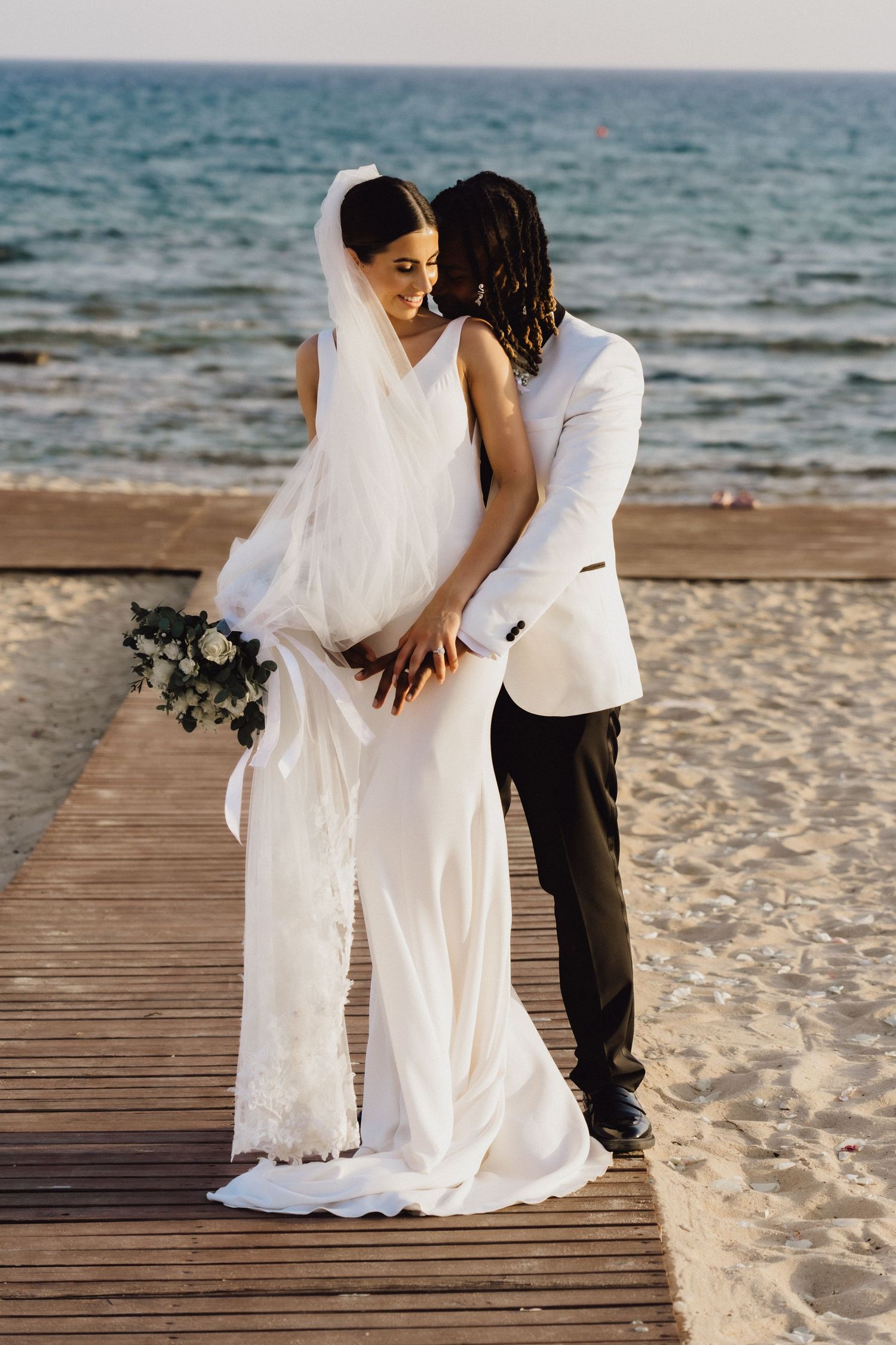 Wedding couple shares intimate moment on wooden boardwalk by Mediterranean beach.