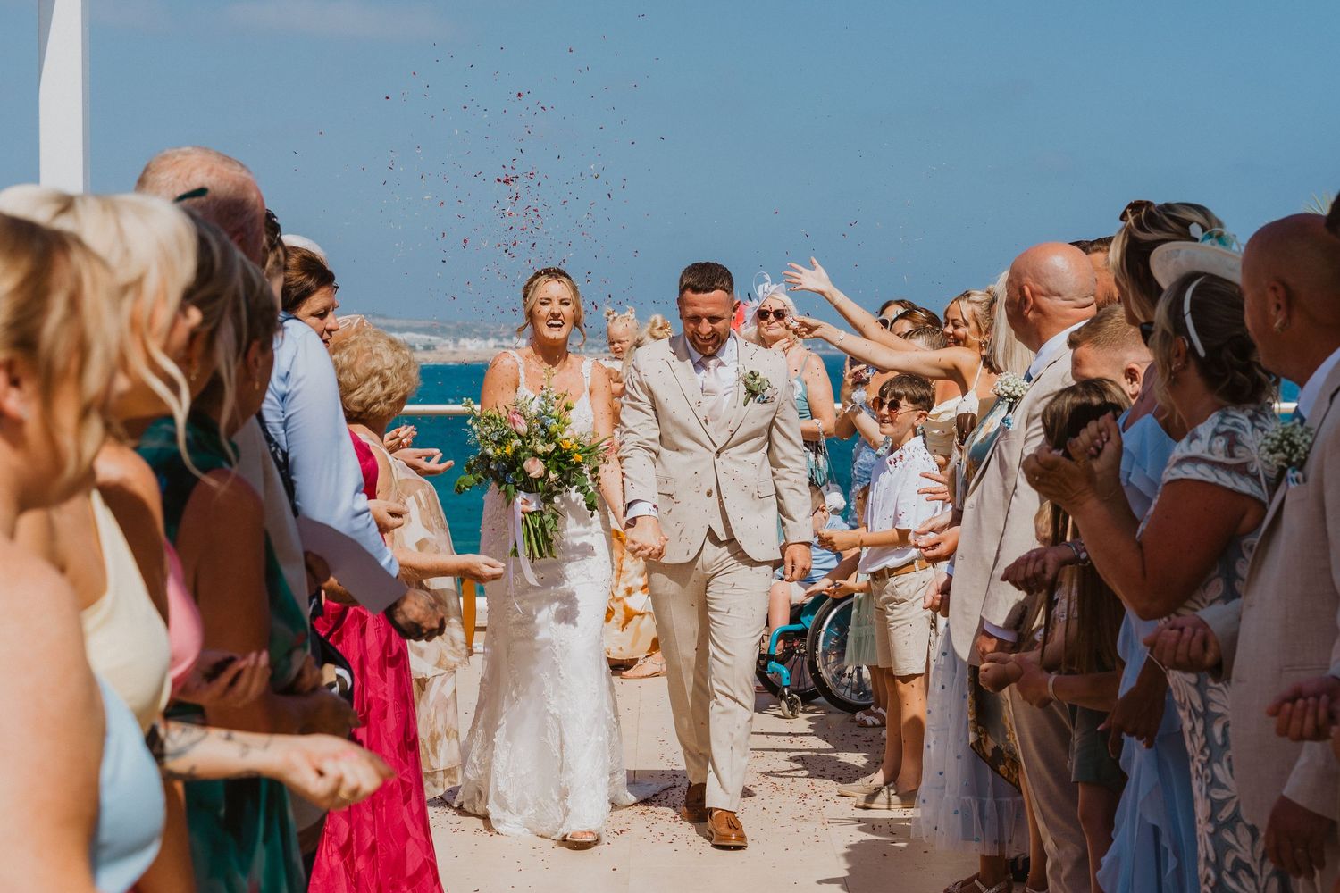 Wedding guests celebrate on a sunny beach by throwing confetti and rose petals in the air during a joyful ceremony.