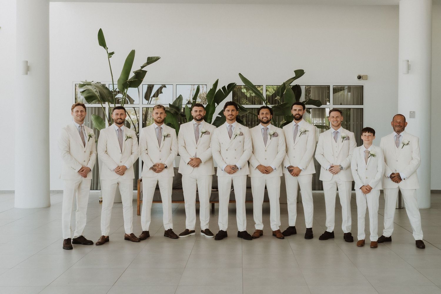 Wedding party dressed in white attire poses together in a line against a light colored wall.