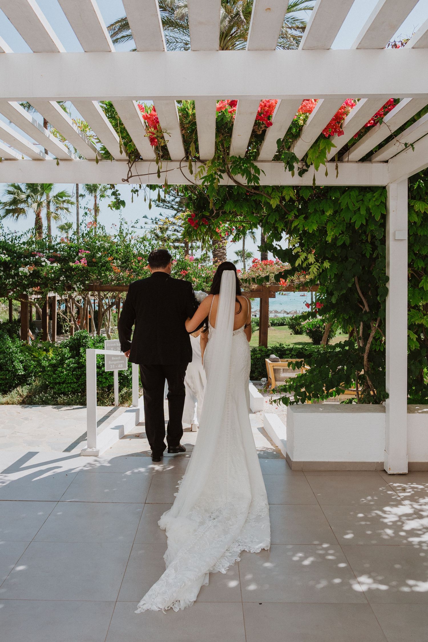 Wedding couple walking under a white pergola decorated with greenery and flowers.