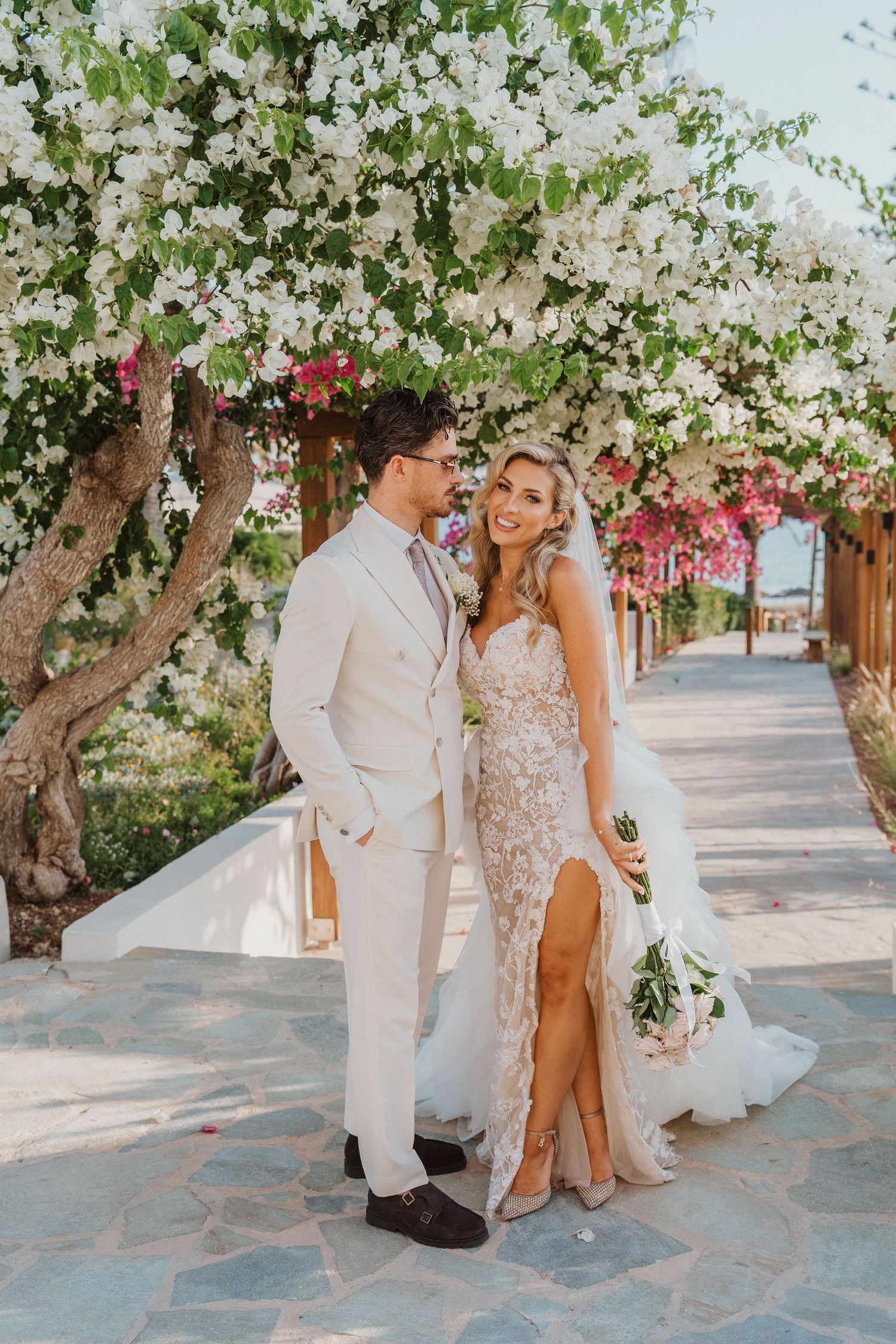 A romantic couple poses under blooming white floral trees on a stone pathway in elegant wedding attire.