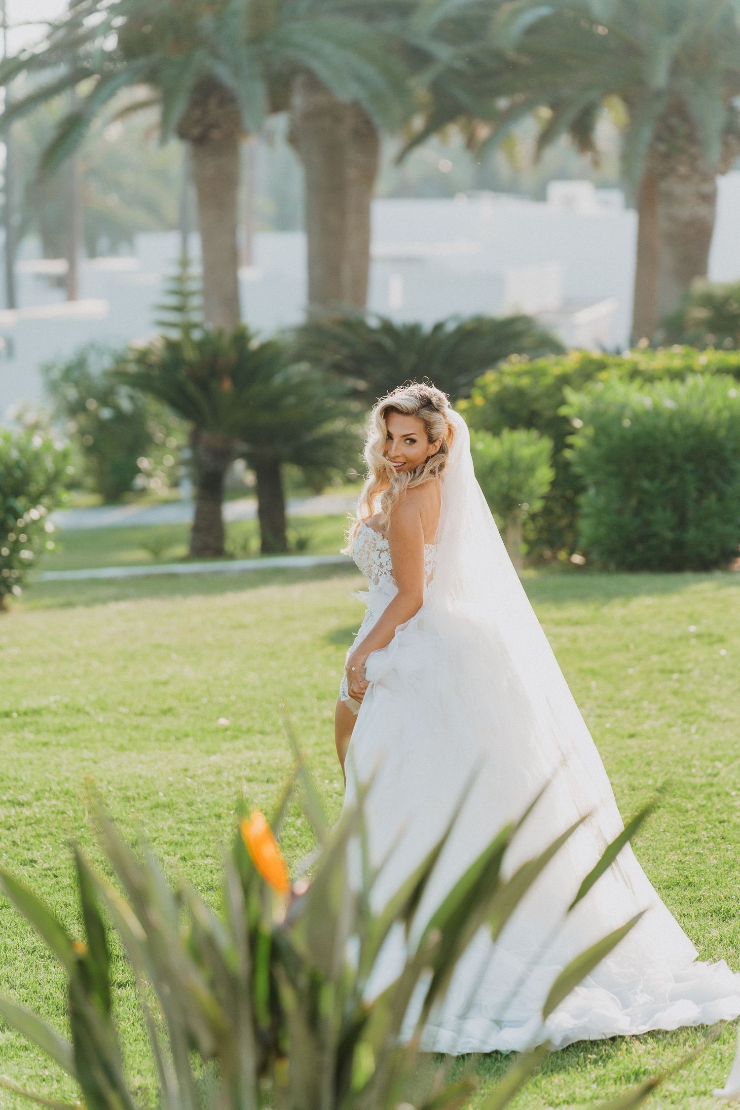A bride in a flowing white wedding dress poses gracefully on a grassy lawn surrounded by tropical palm trees.