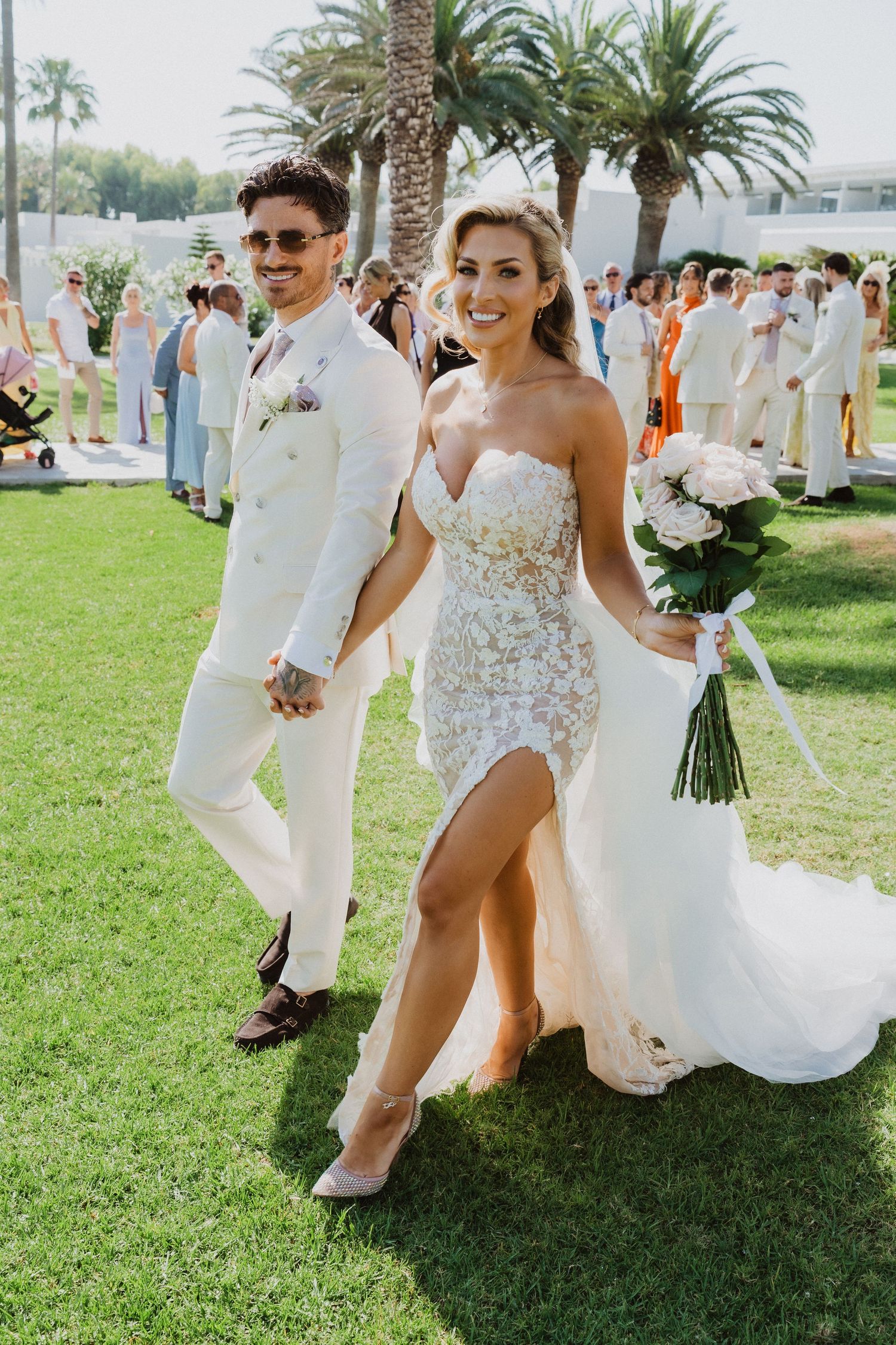 Wedding party walking and celebrating on lush green lawn with palm trees in background during outdoor ceremony.
